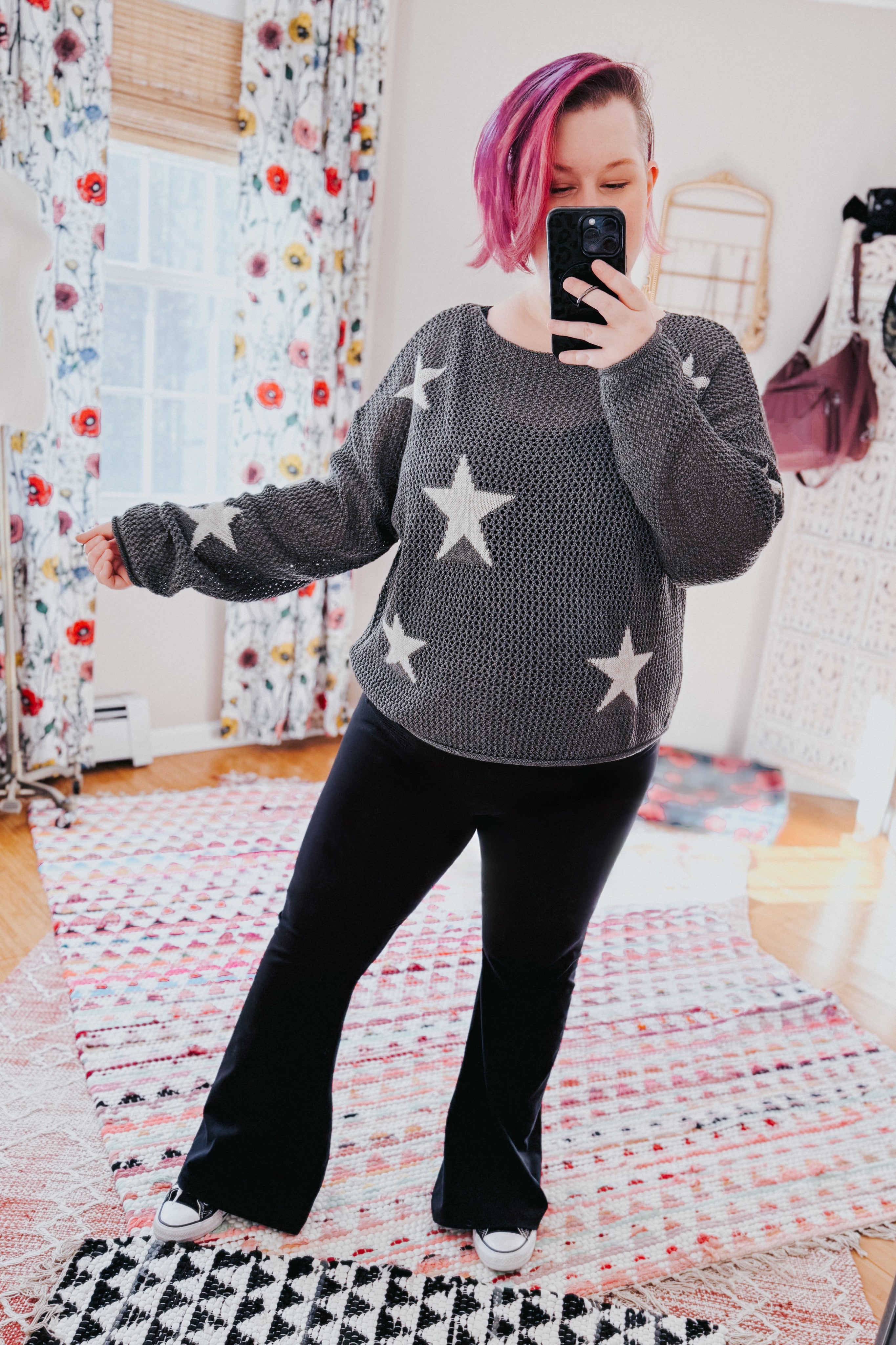 Woman with pink hair wearing charcoal mesh sweater with star patterns paired with black flared pants taking selfie indoors