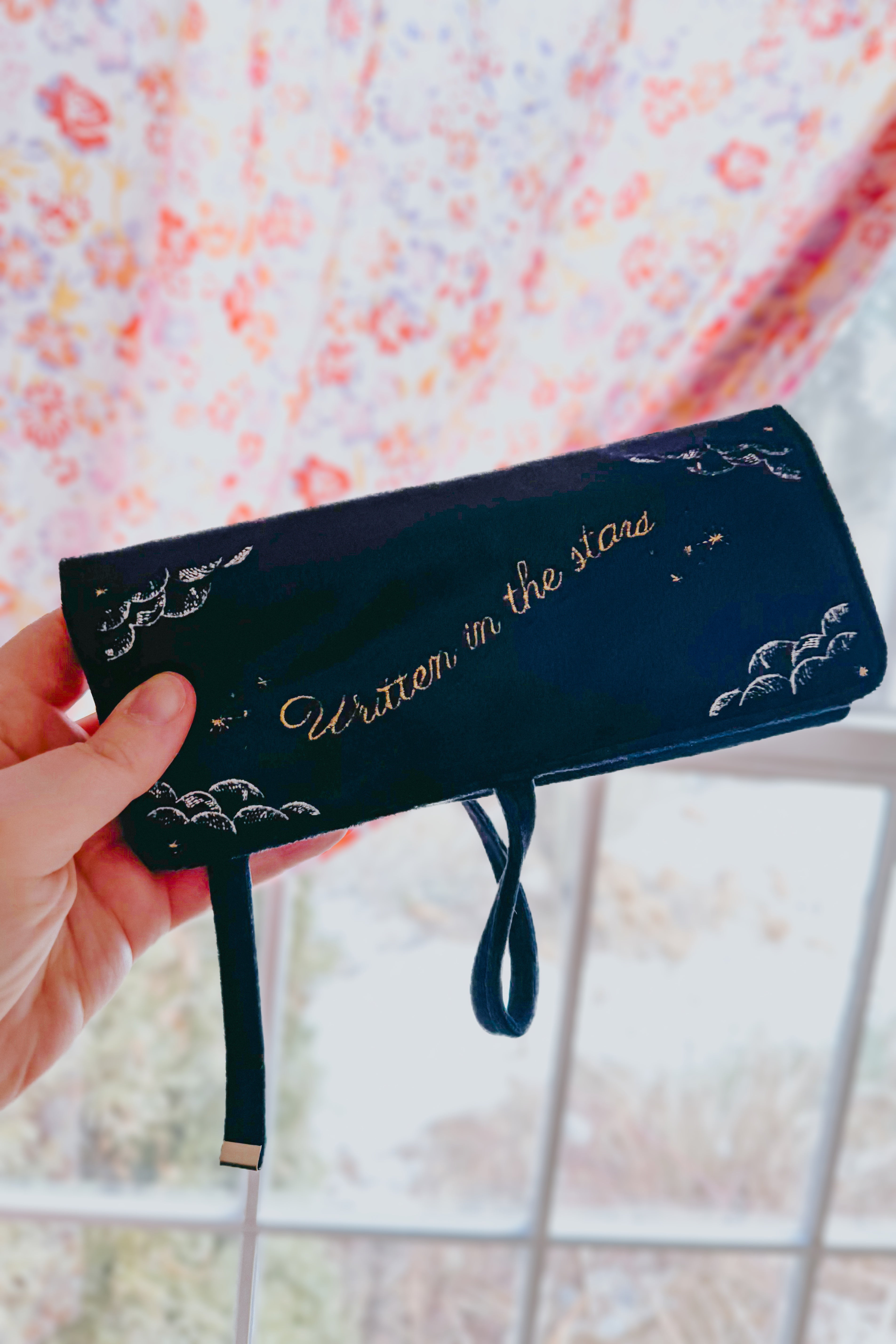 Hand holding navy velvet jewelry roll embroidered with "Written in the stars" and celestial designs indoors by window