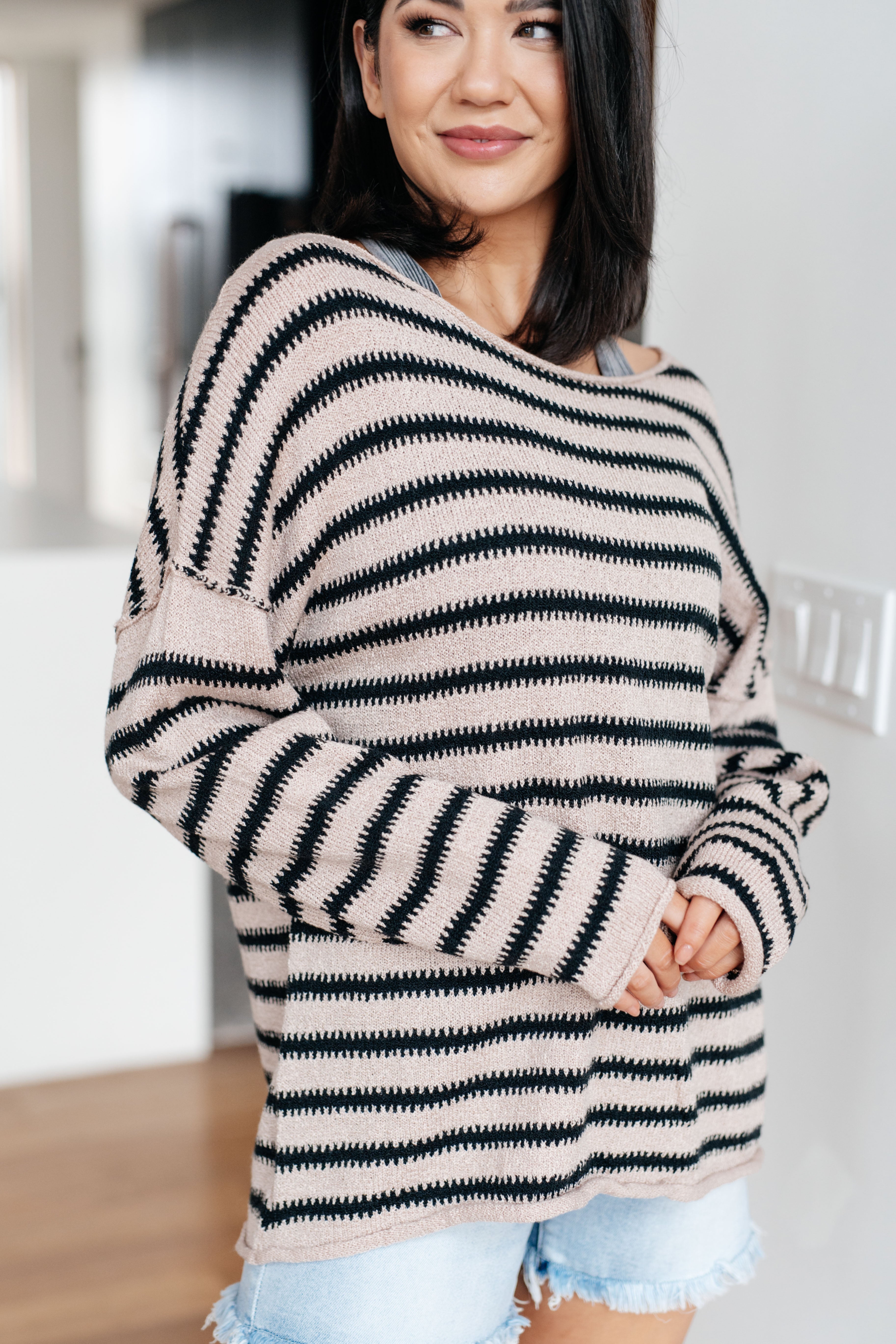 Woman wearing a taupe sweater with black stripes and denim shorts indoors.