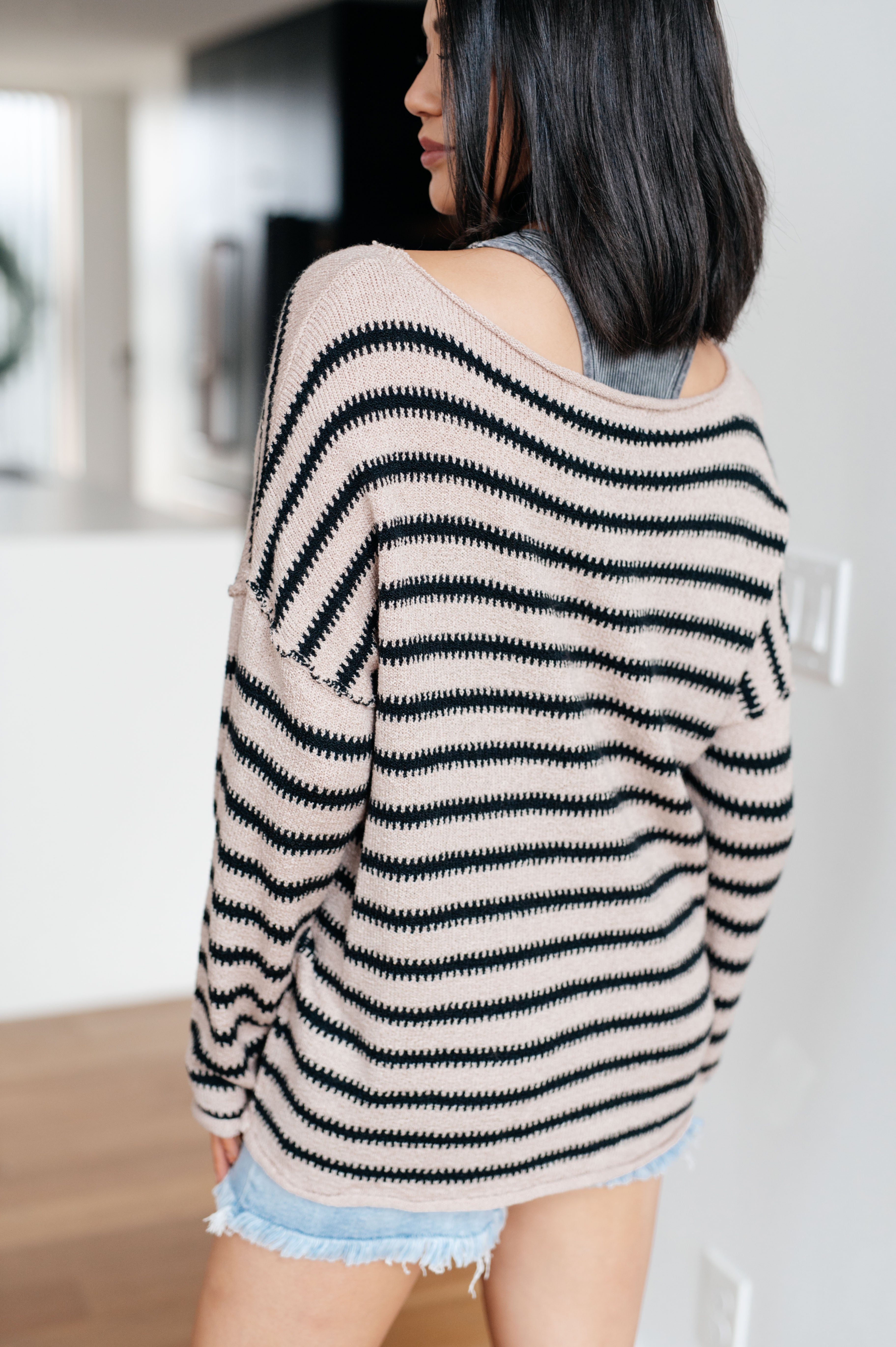 Woman wearing a taupe sweater with black stripes and denim shorts indoors.
