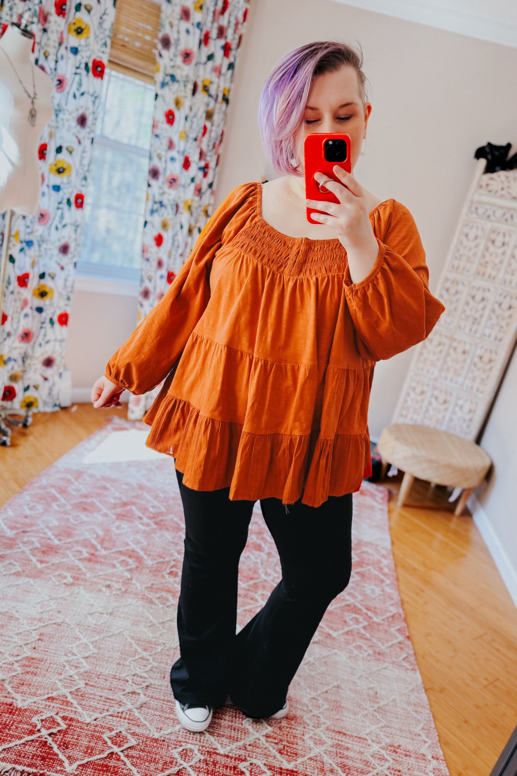 Woman with purple hair wearing rust ruched babydoll top and black pants taking mirror selfie indoors