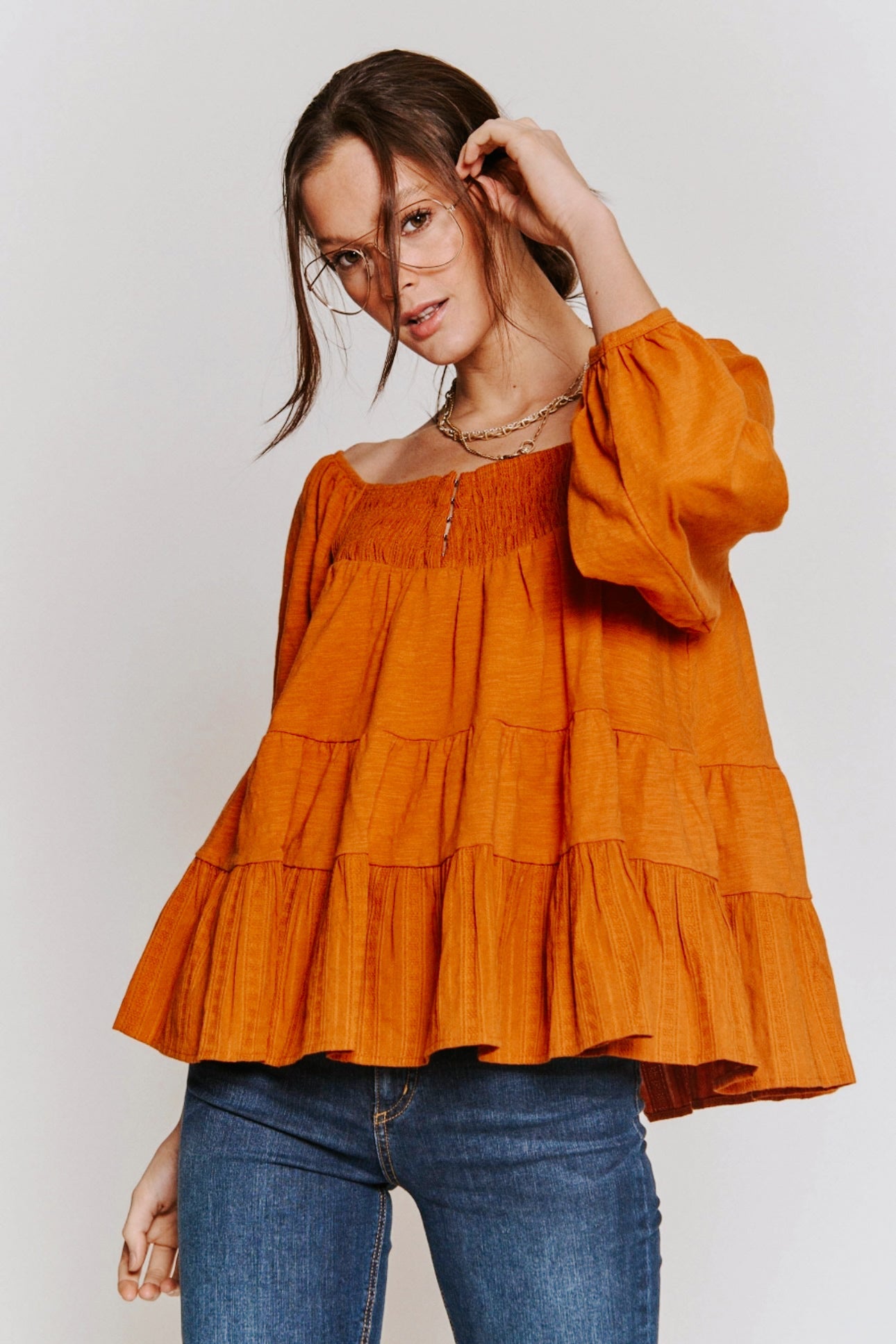 Woman wearing rust-colored ruched babydoll top with puff sleeves and layered hem paired with blue jeans