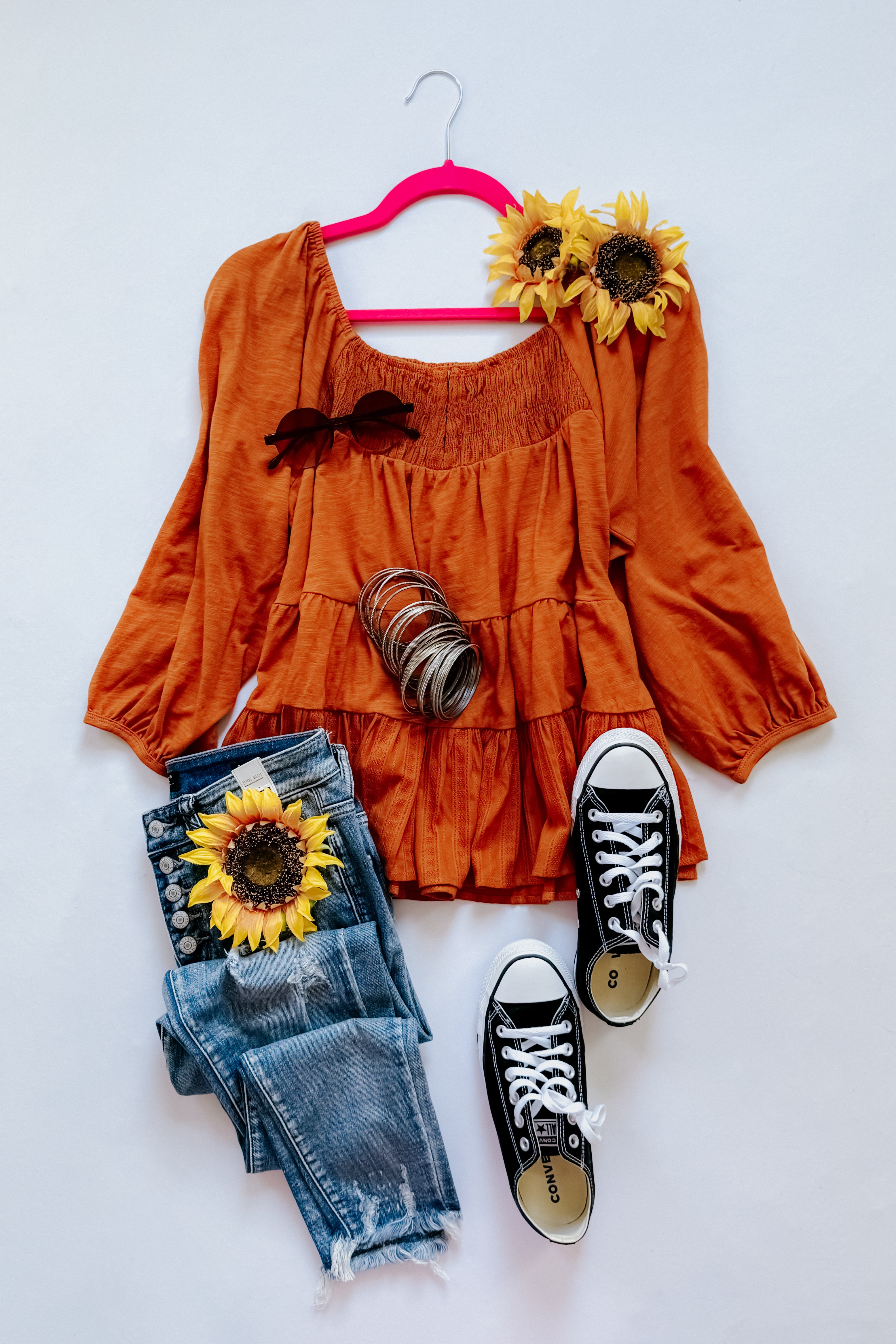 Rust-colored ruched babydoll top paired with distressed blue jeans, black sneakers, bracelets, and sunflower accents on white background