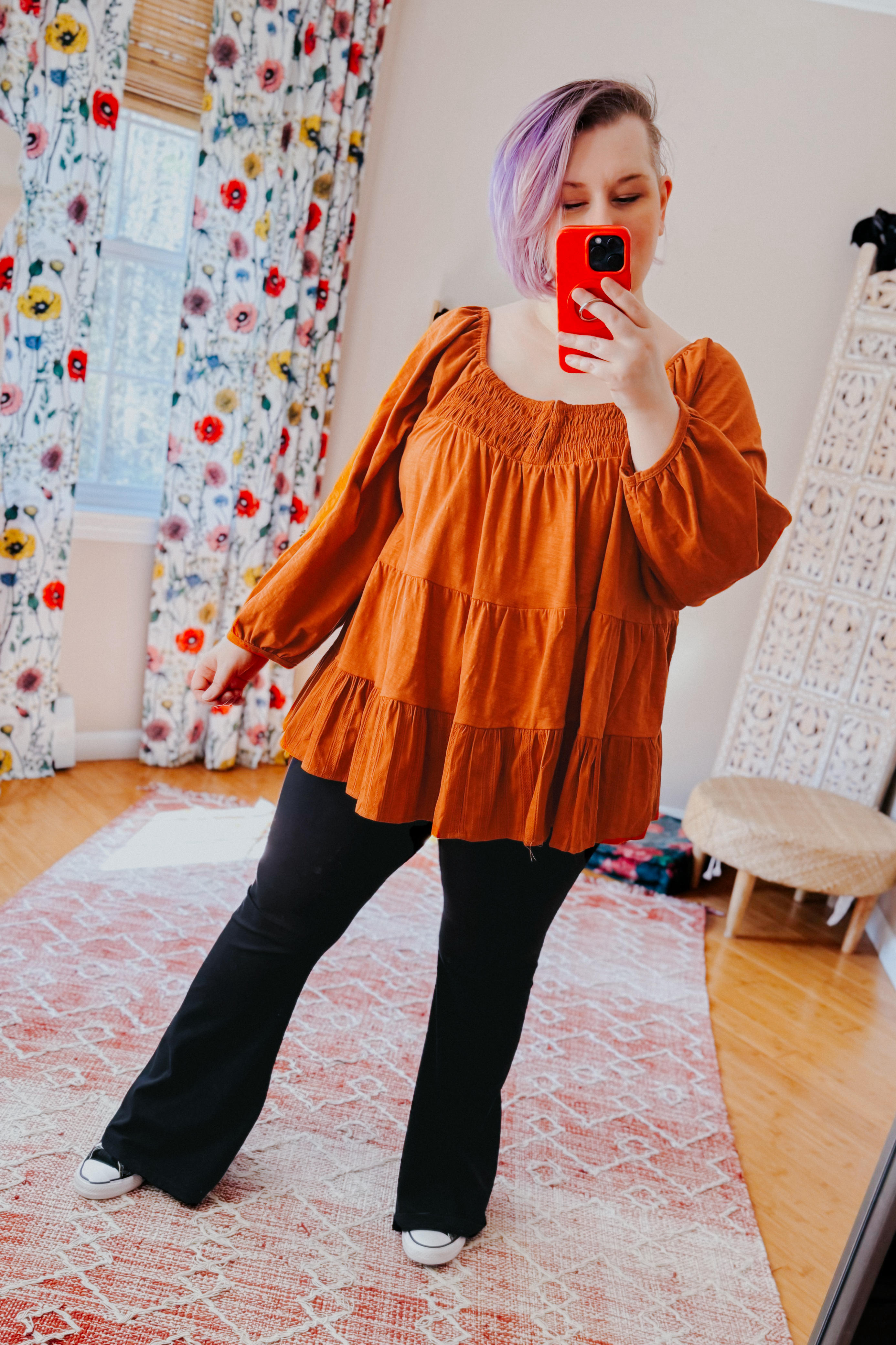 Woman with purple hair wearing rust ruched babydoll top and black pants taking mirror selfie in bright room