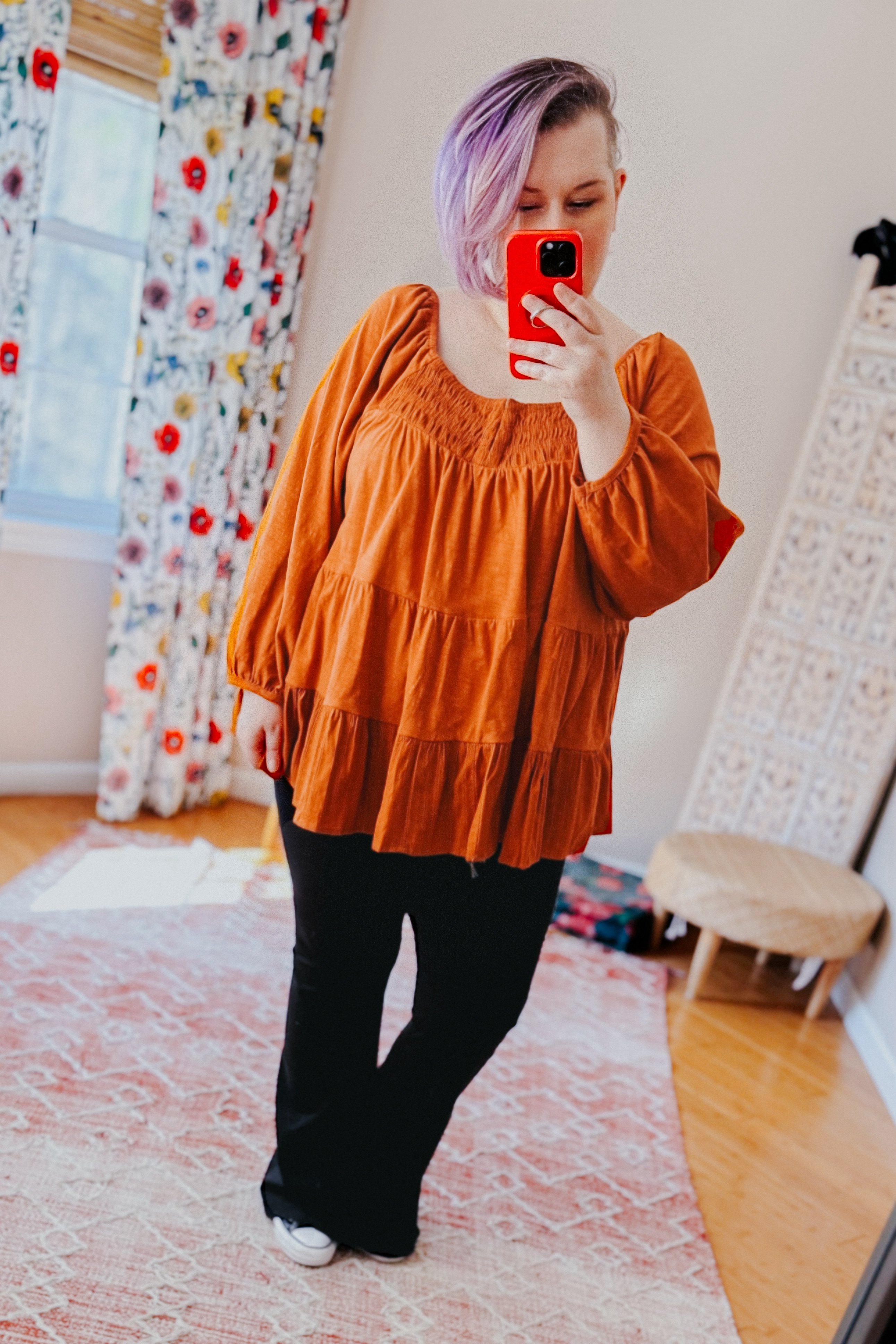 Woman taking mirror selfie wearing rust-colored ruched babydoll top with black pants in cozy room with floral curtains
