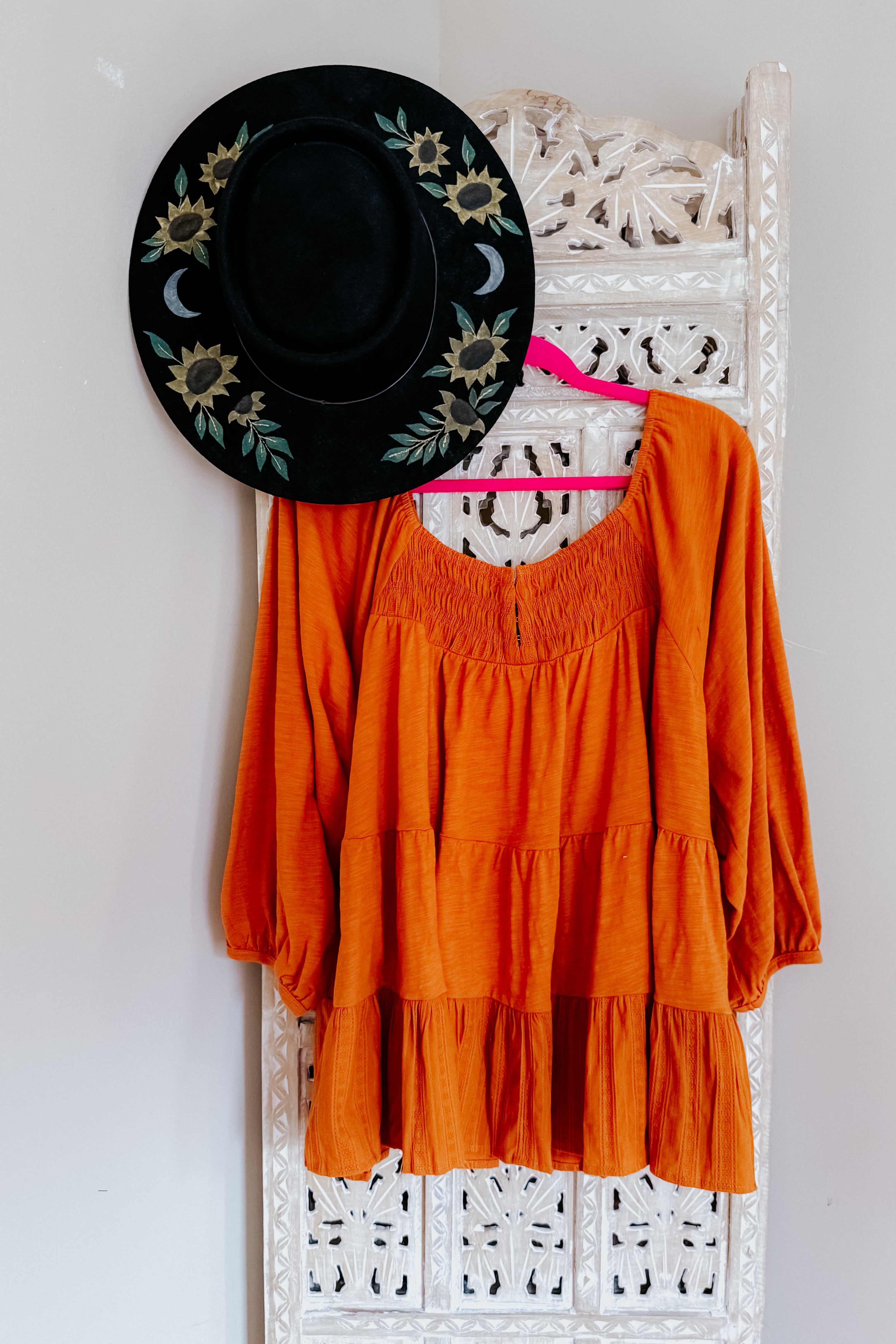 Rust-colored ruched babydoll top with long sleeves hanging on carved white wood panel next to black floral hat