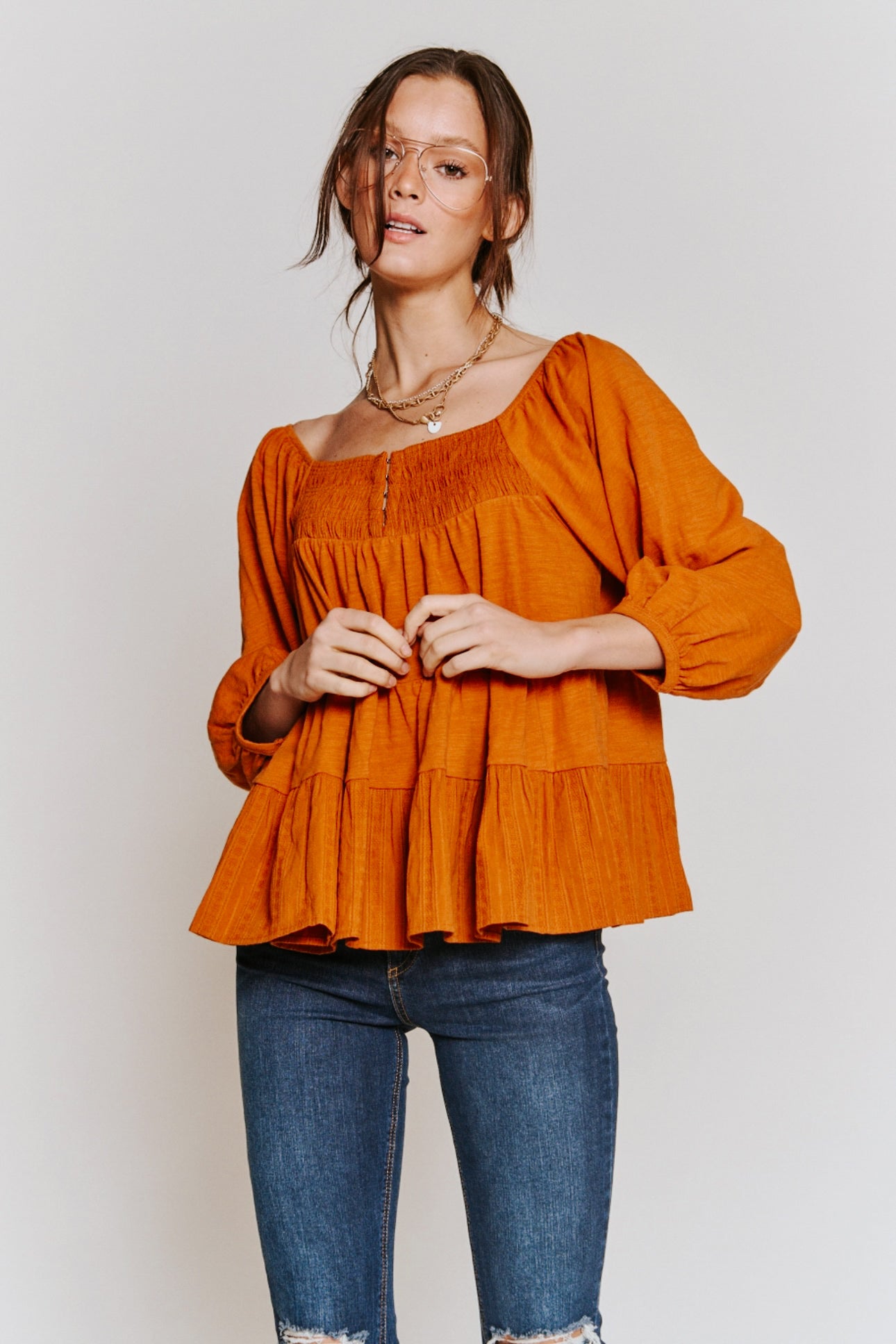 Woman wearing rust-colored ruched babydoll top with puff sleeves paired with blue distressed jeans on neutral background