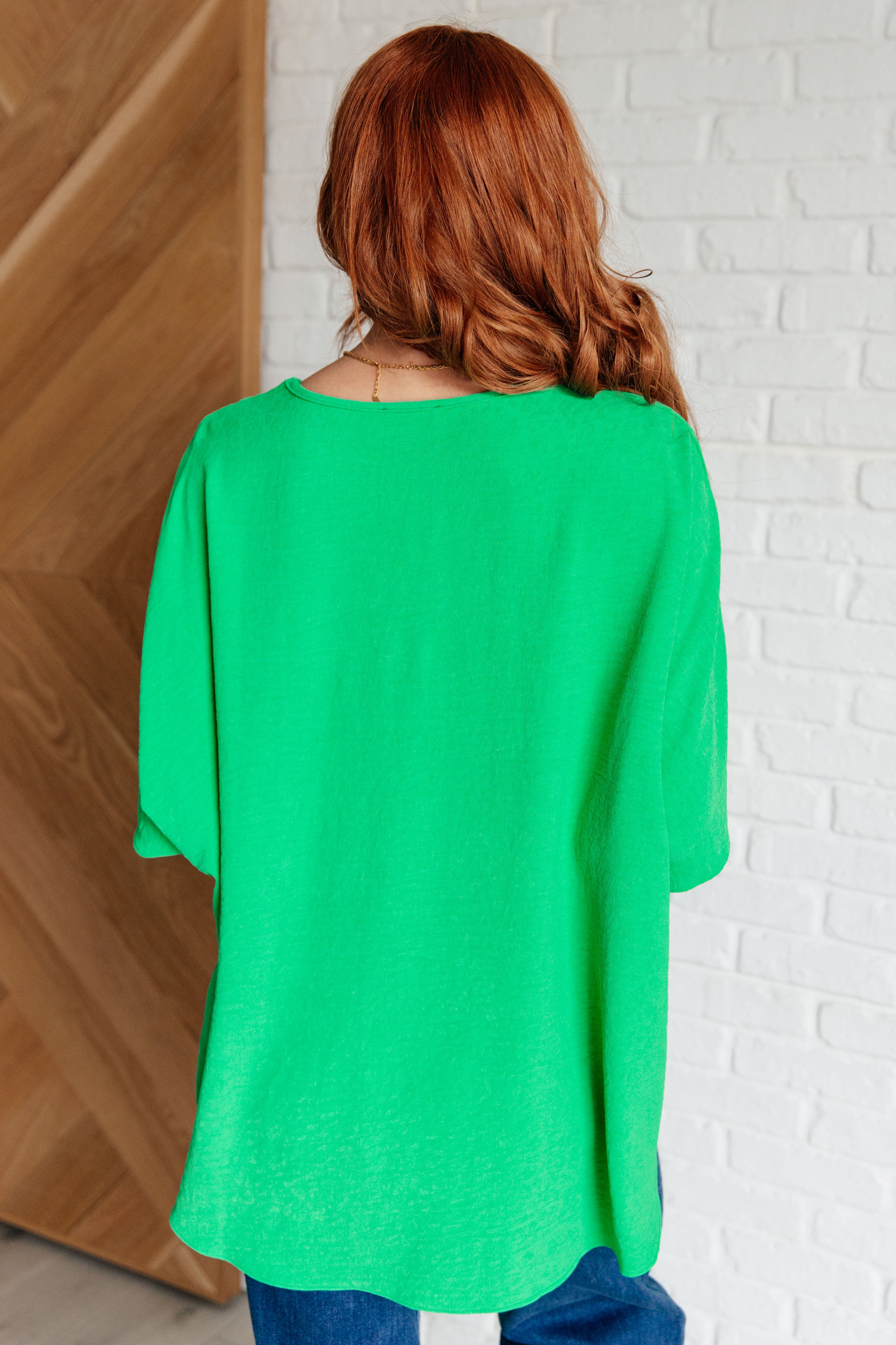 Woman with red hair wearing a loose bright green V-neck top standing indoors against white brick and wood walls