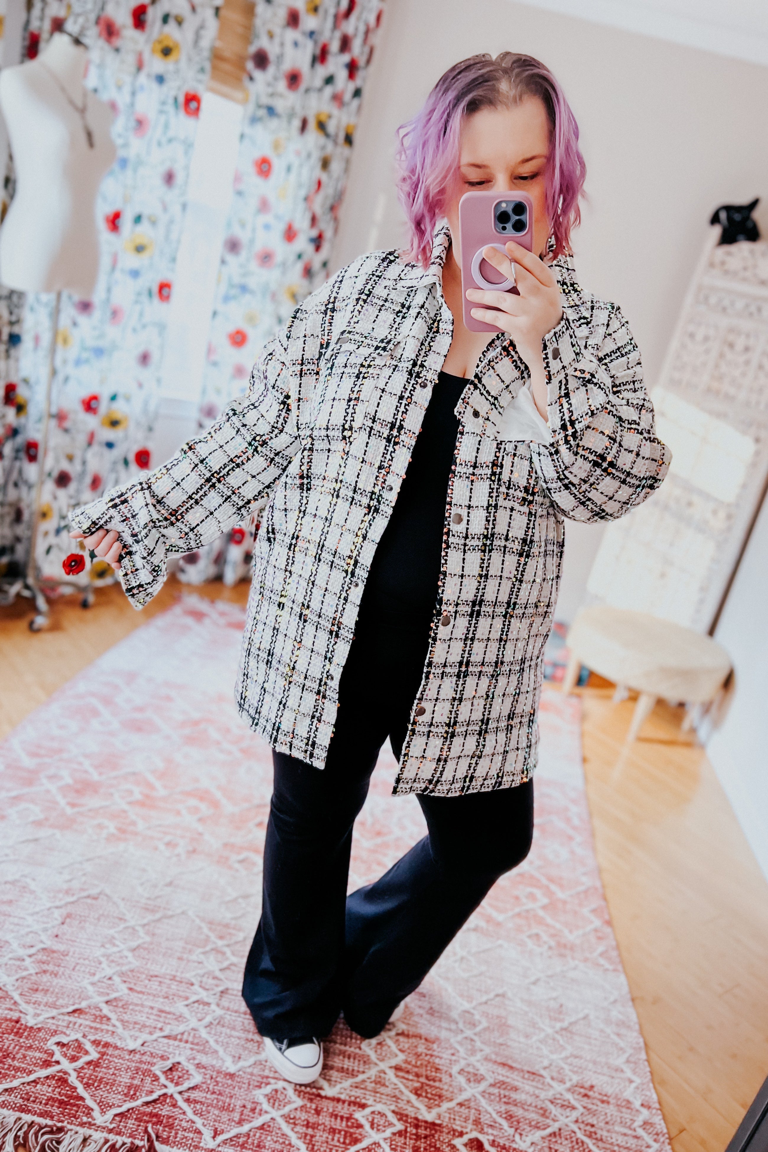 Woman with purple hair wearing white plaid sequin shacket over black top and pants taking mirror selfie indoors