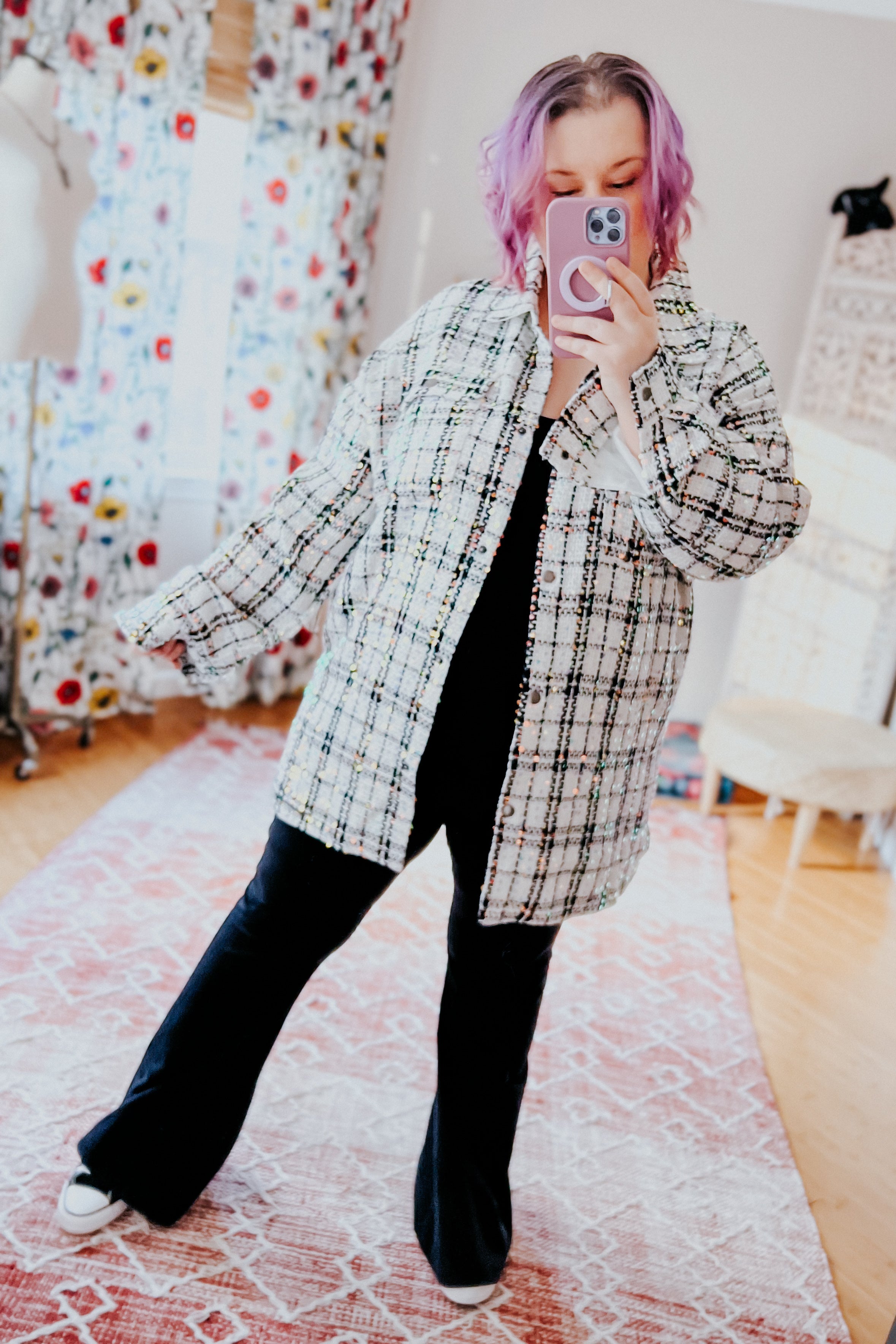 Woman with purple hair wearing white plaid sequin shacket over black outfit taking mirror selfie indoors