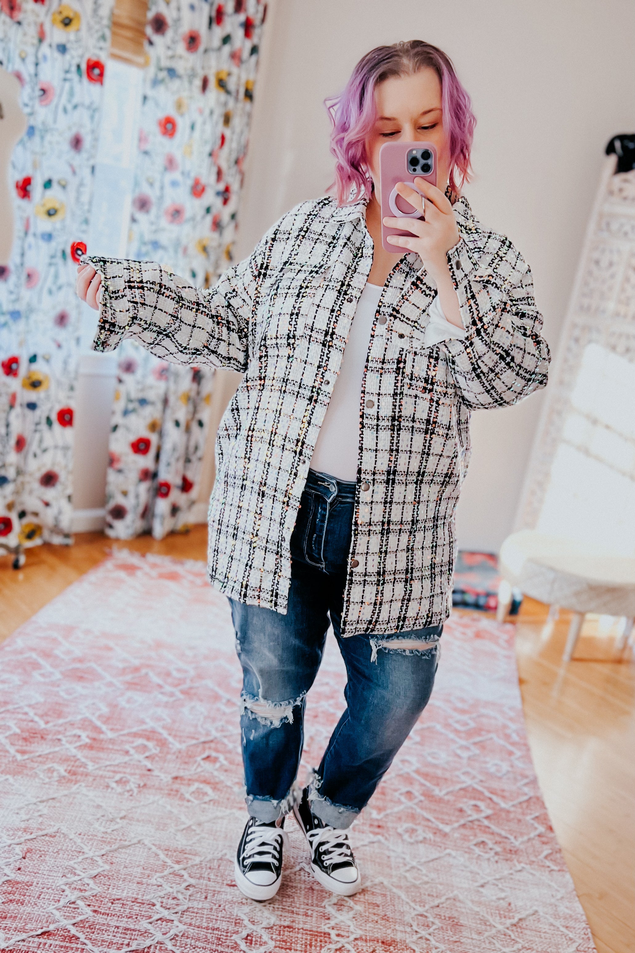 Woman with pastel purple hair wearing white plaid sequin shacket over white top and ripped dark jeans taking mirror selfie