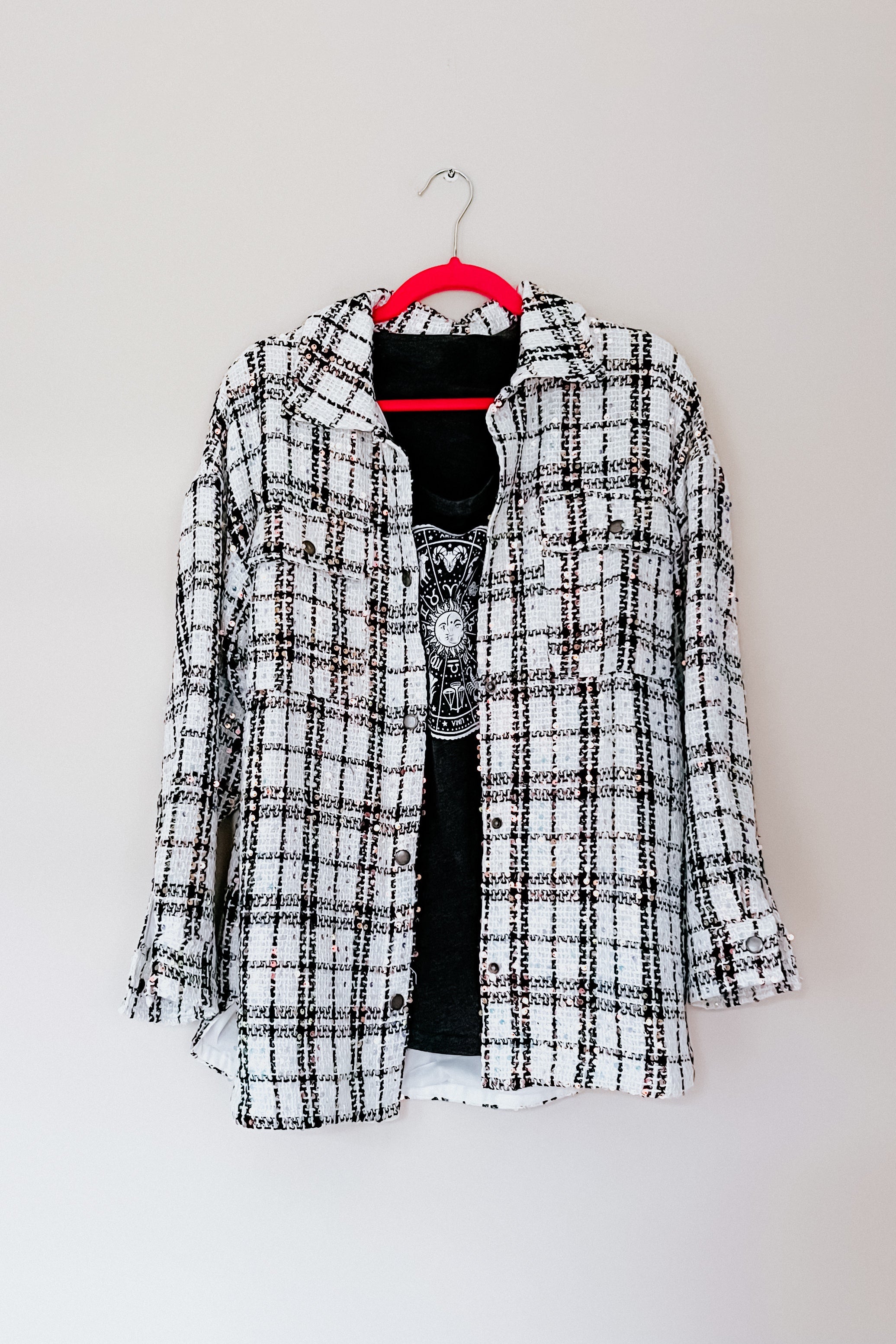 White plaid sequin shacket layered over black graphic top hanging on bright pink hanger against white wall