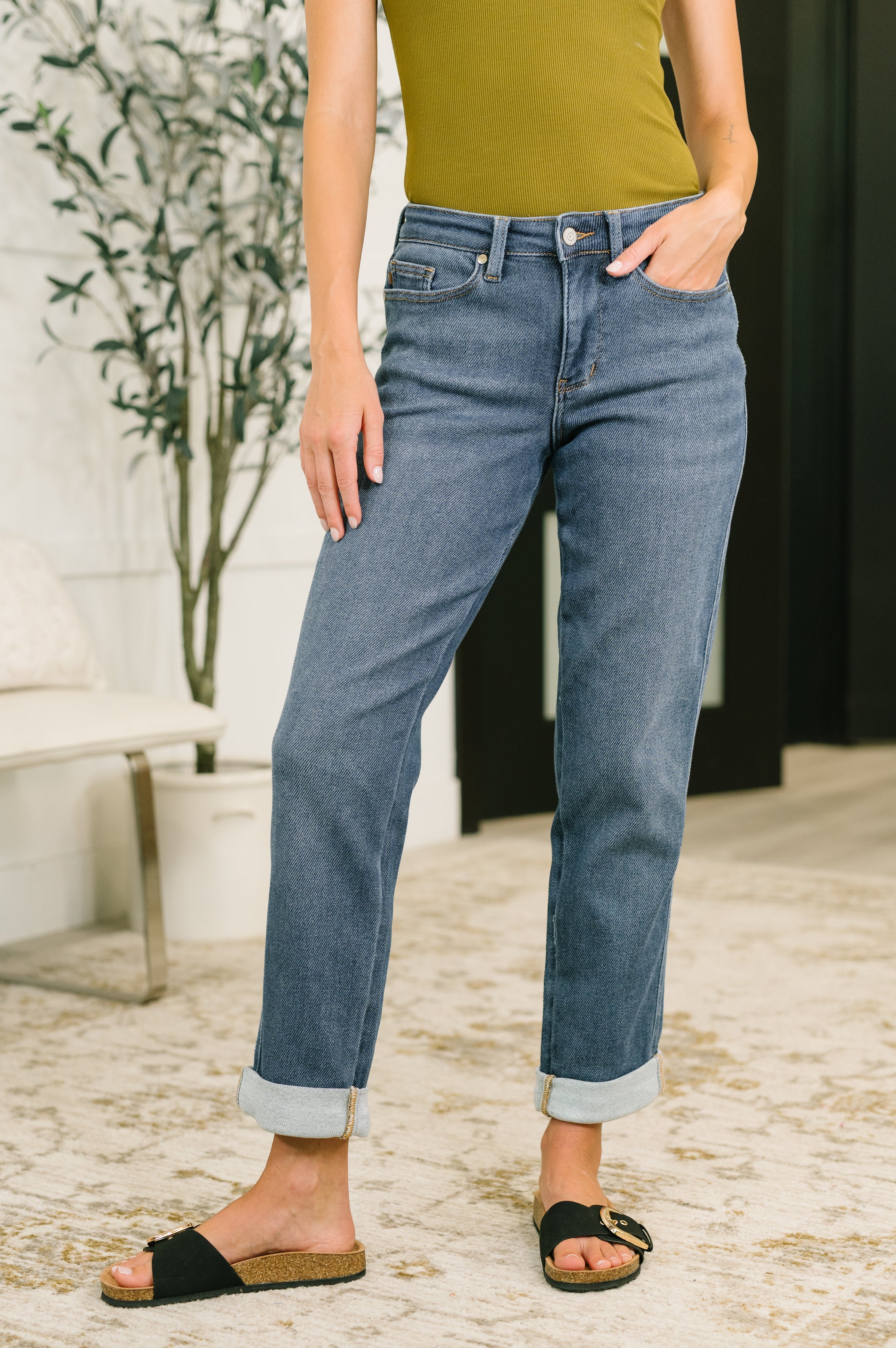 Woman wearing mid-rise blue denim boyfriend jeans with rolled cuffs and black slide sandals indoors