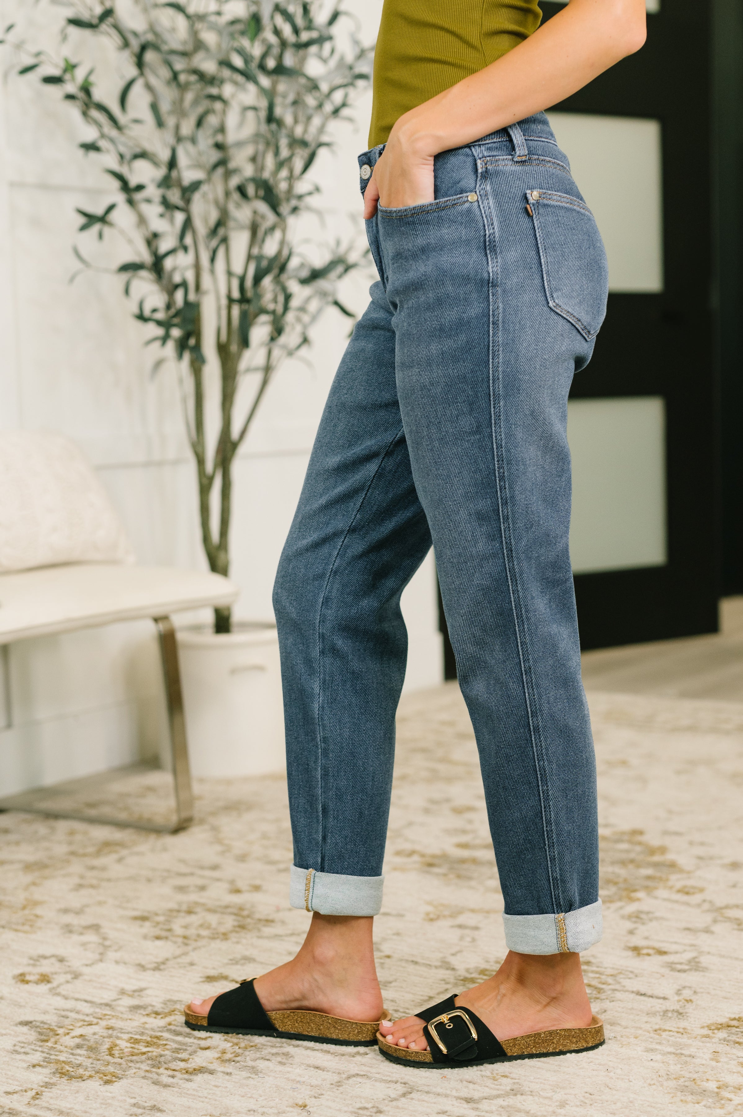 Side view of relaxed mid rise blue denim jeans with cuffed hems worn with olive top and black slide sandals indoors
