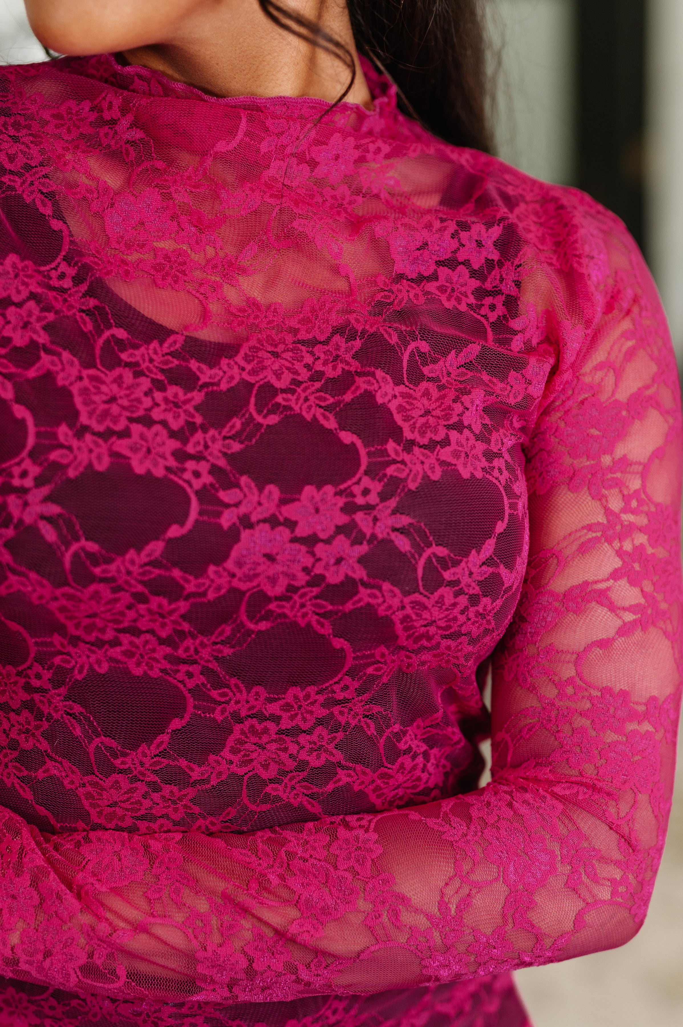 Close-up of a sheer fuchsia lace top with floral pattern worn by person with dark hair in soft lighting