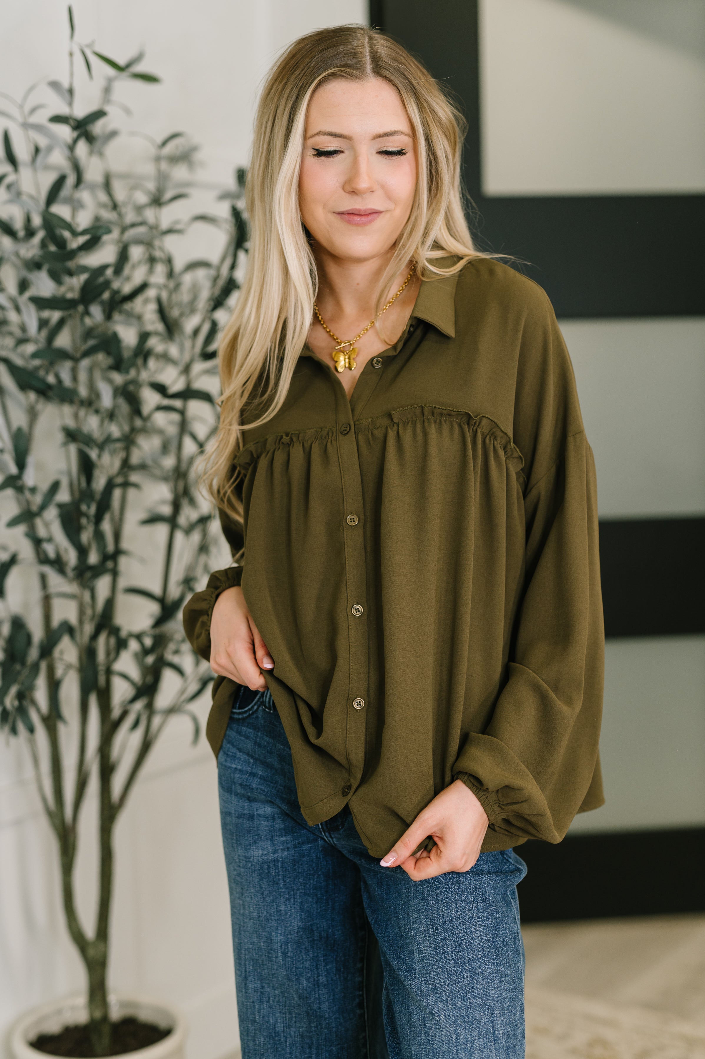 Woman wearing olive button-down blouse with gathered yoke and balloon sleeves paired with blue jeans indoors
