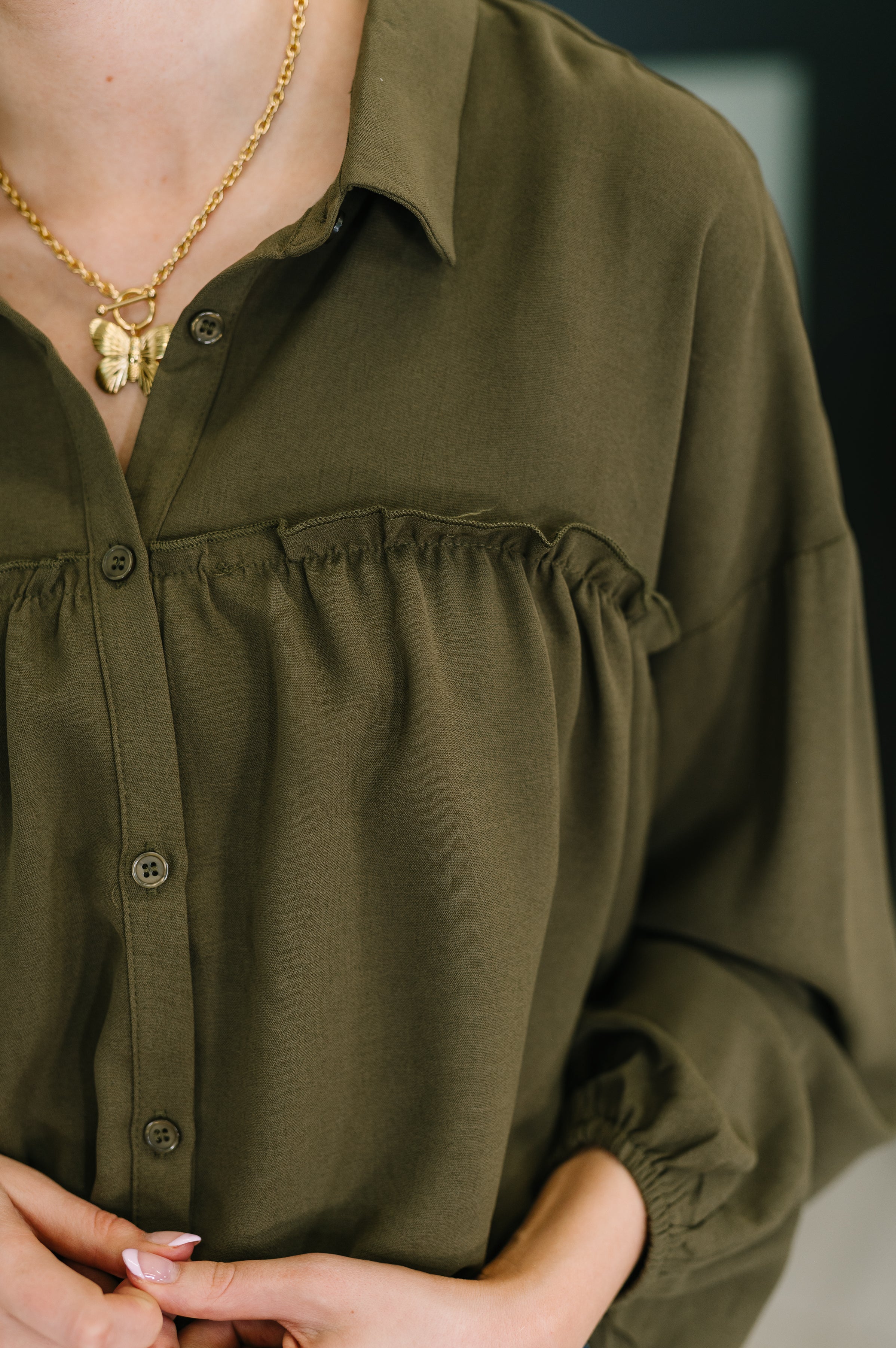 Close-up of olive button-down blouse with ruffle detail across chest and gold butterfly pendant necklace