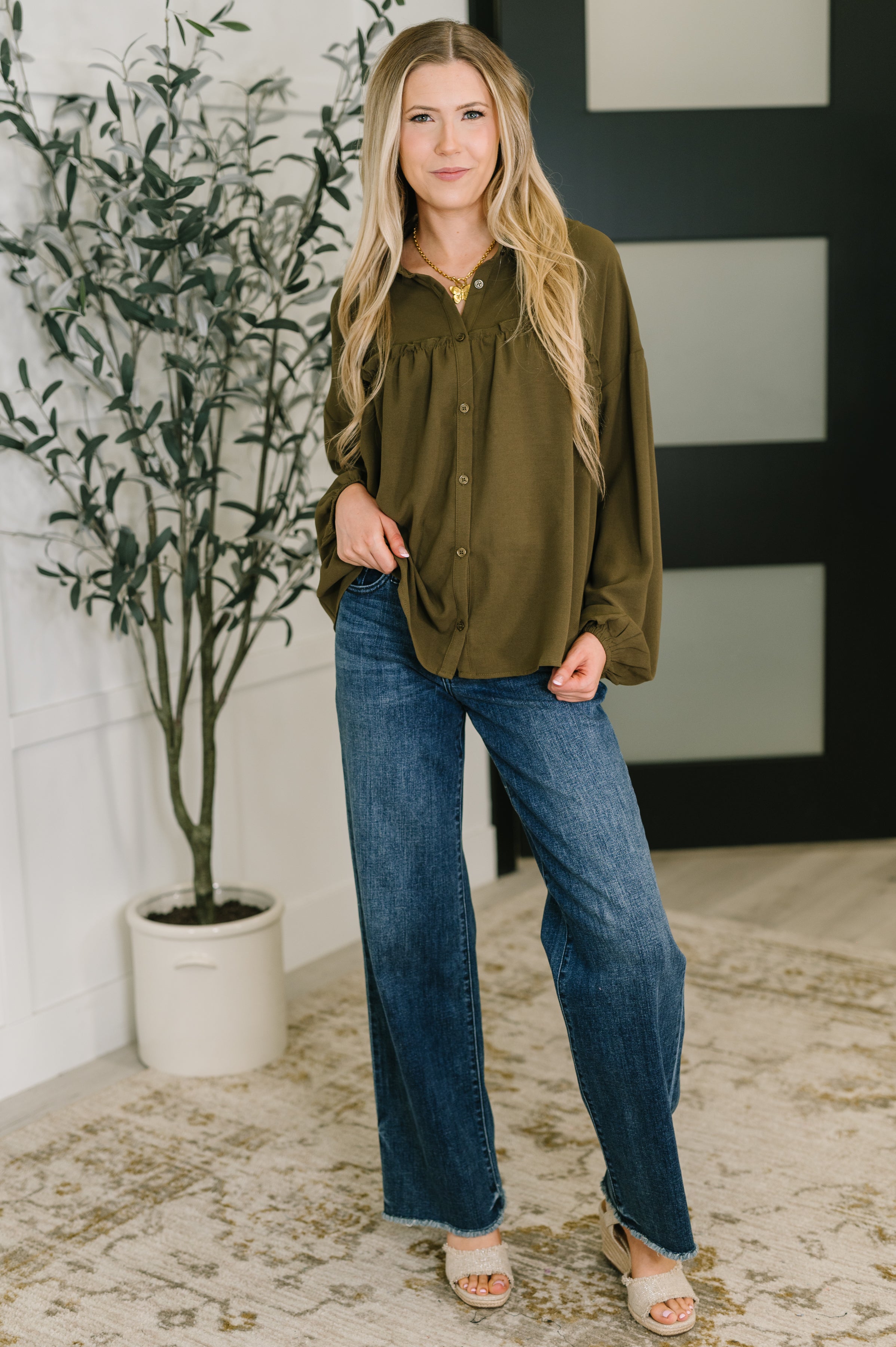 Woman wearing olive button-down blouse paired with high-waisted wide-leg jeans and sandals indoors near plant