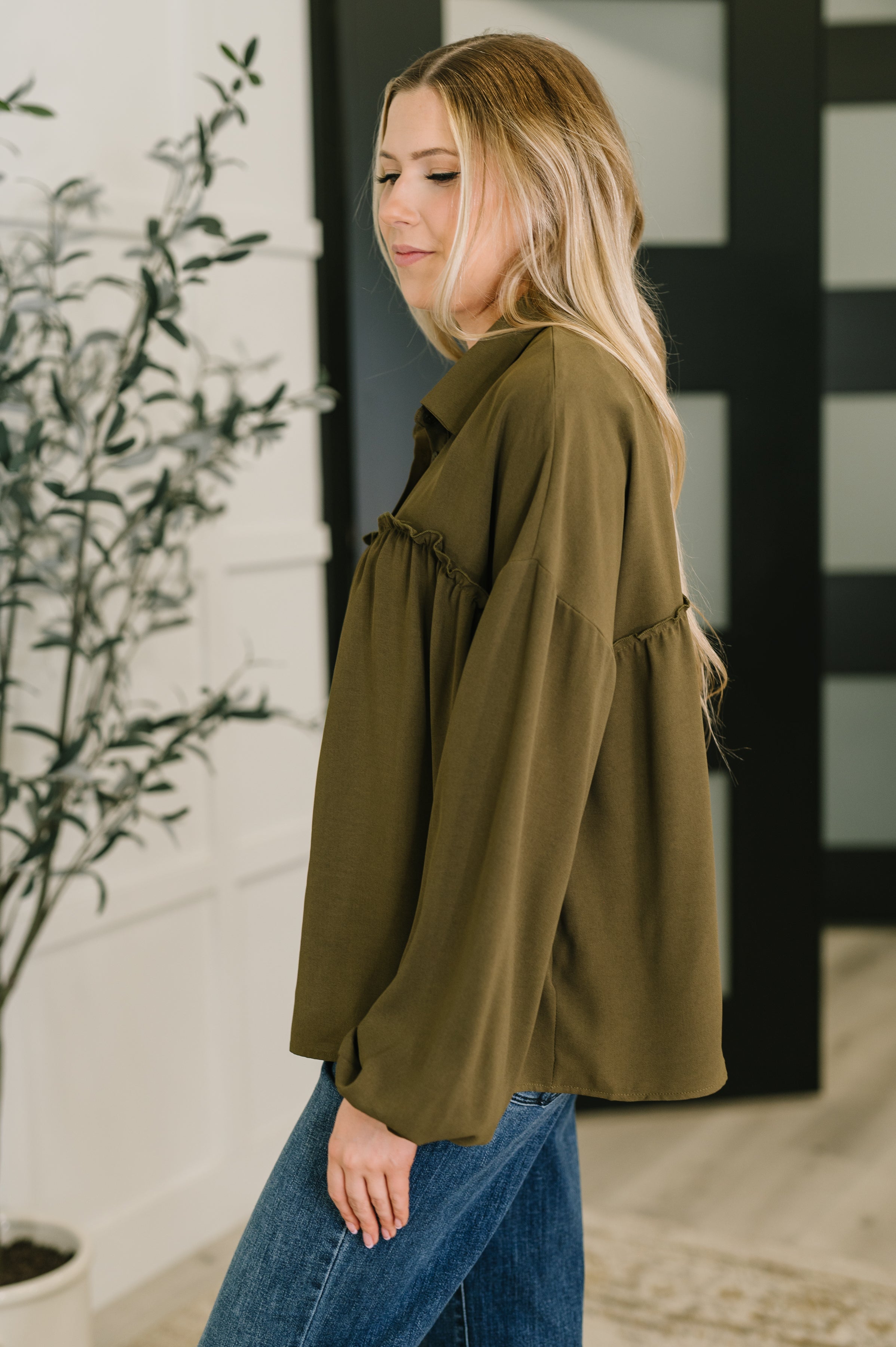 Woman wearing loose olive button down blouse with ruffle detail paired with blue jeans indoors by plant