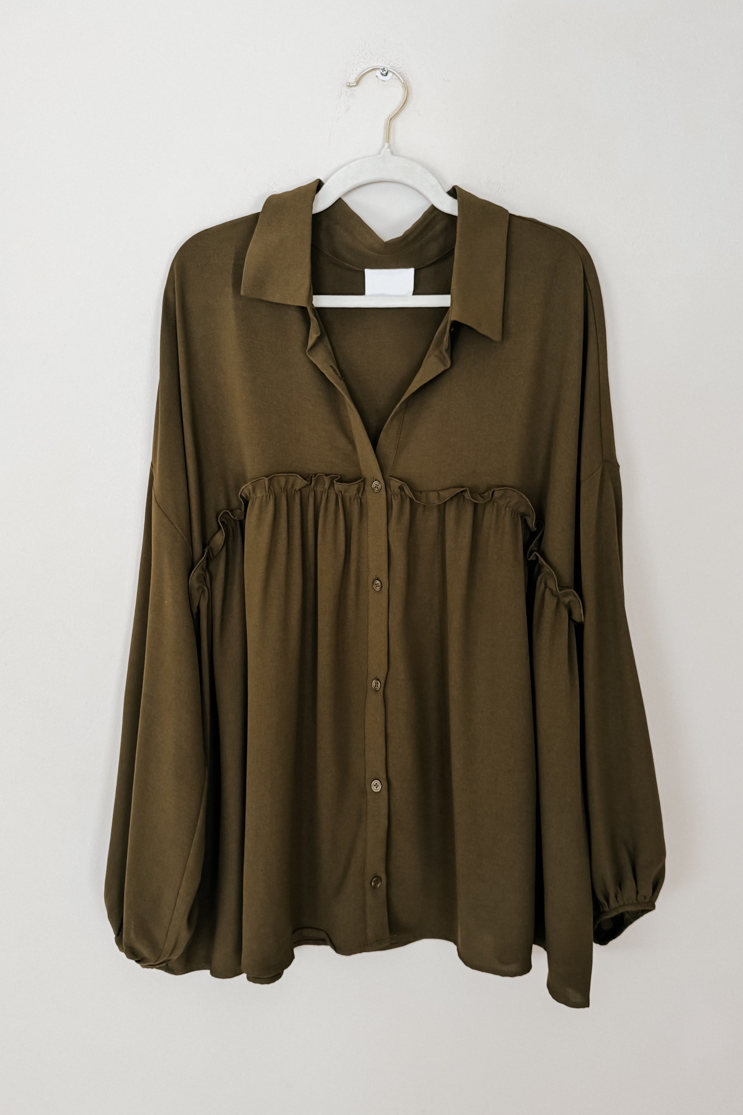 Olive button-down blouse with gathered detailing under bust and loose long sleeves hanging on white hanger against plain wall
