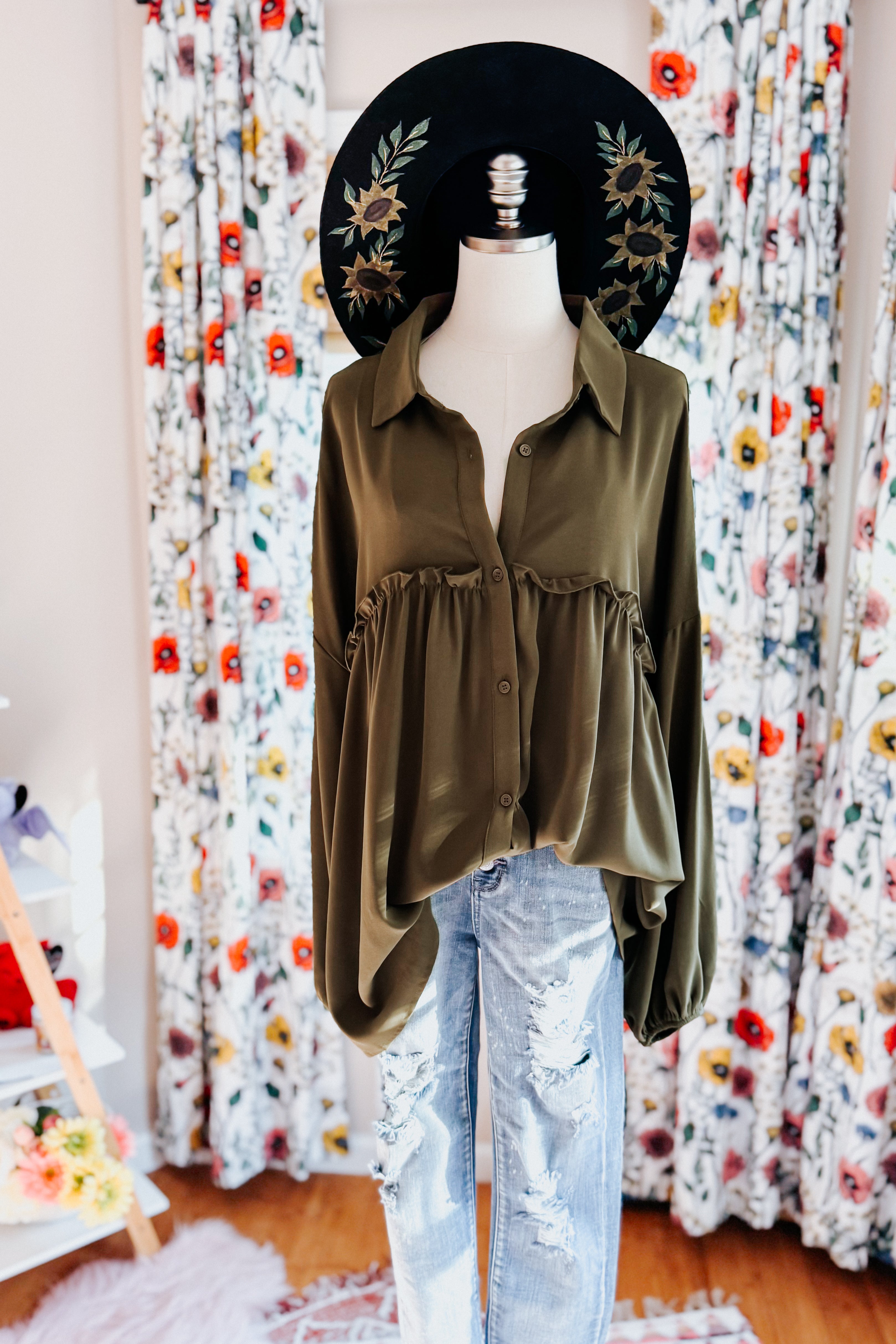 Olive green button-down blouse with ruffle detail paired with distressed light-wash jeans and floral embroidered black hat on mannequin