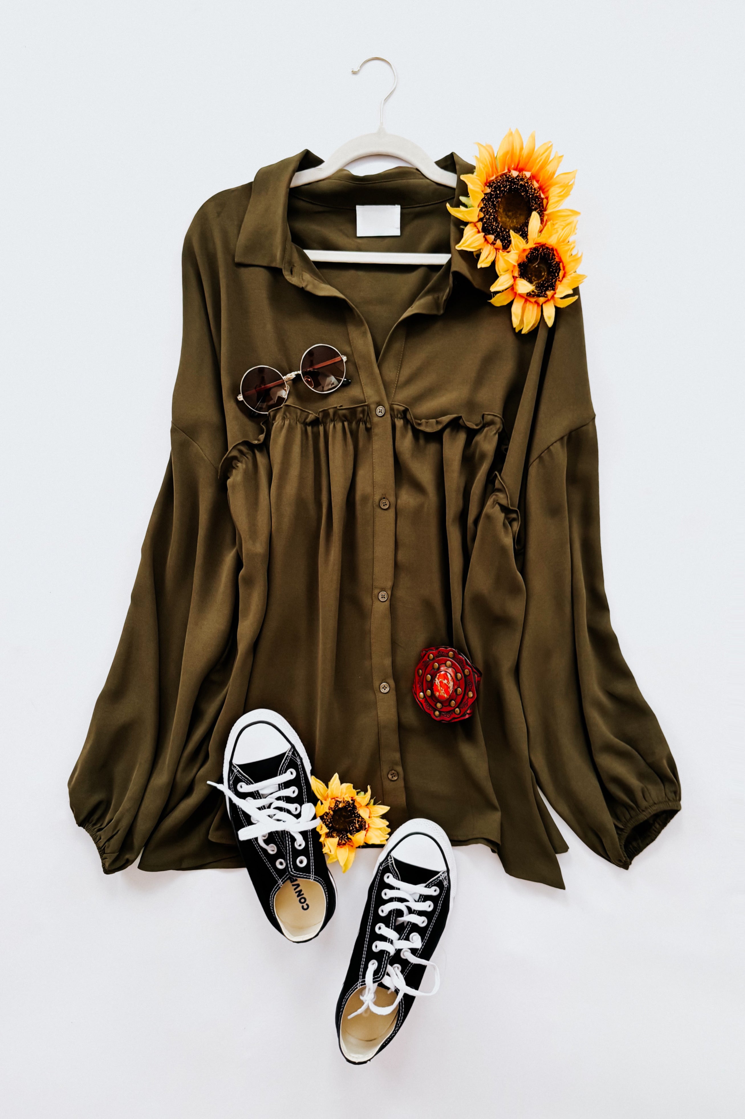 Olive button-down blouse styled with round sunglasses, black sneakers, and vibrant sunflower accents on white background