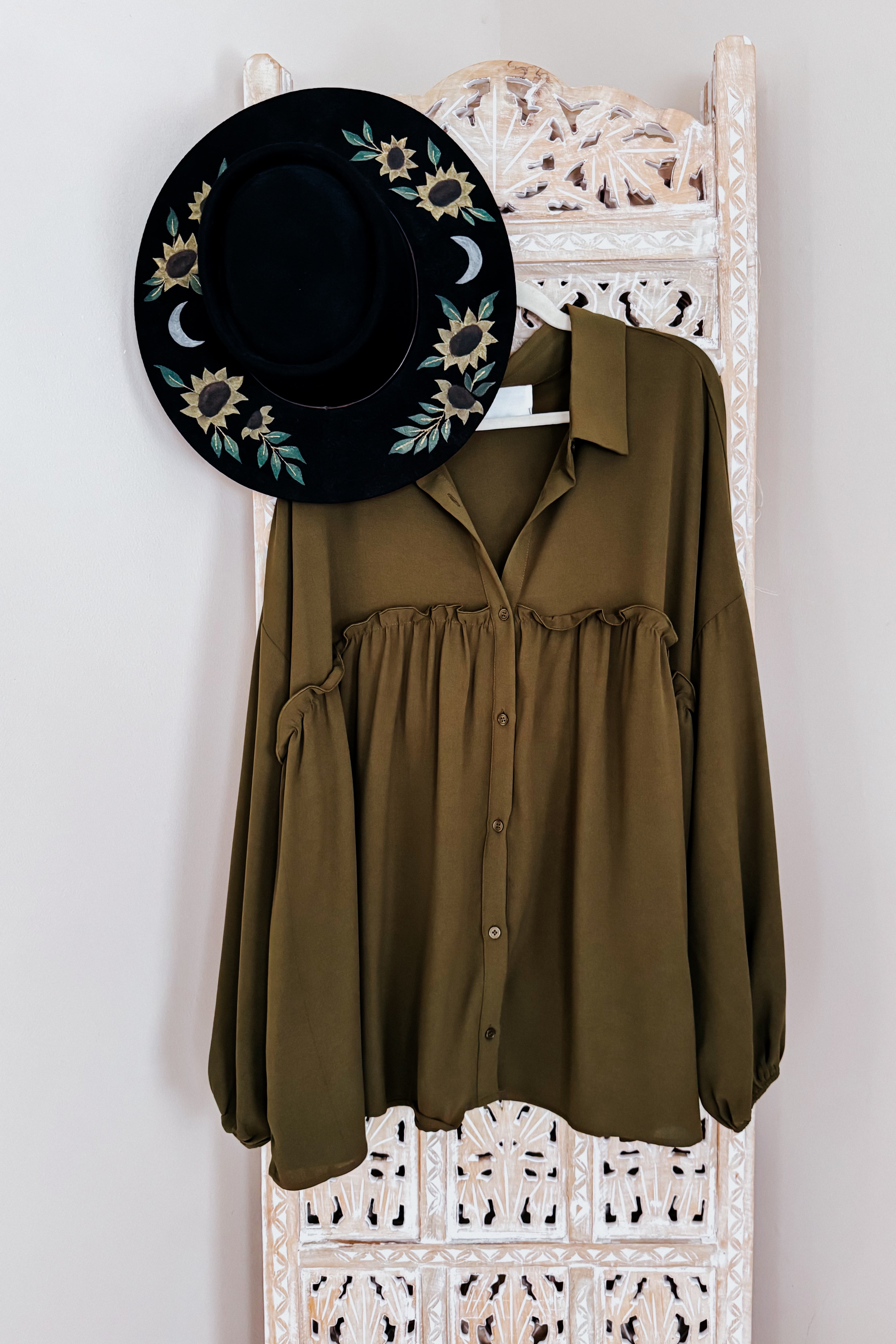 Olive button-down blouse with gathered detailing hanging on carved wooden screen next to black hat with sunflowers and moons