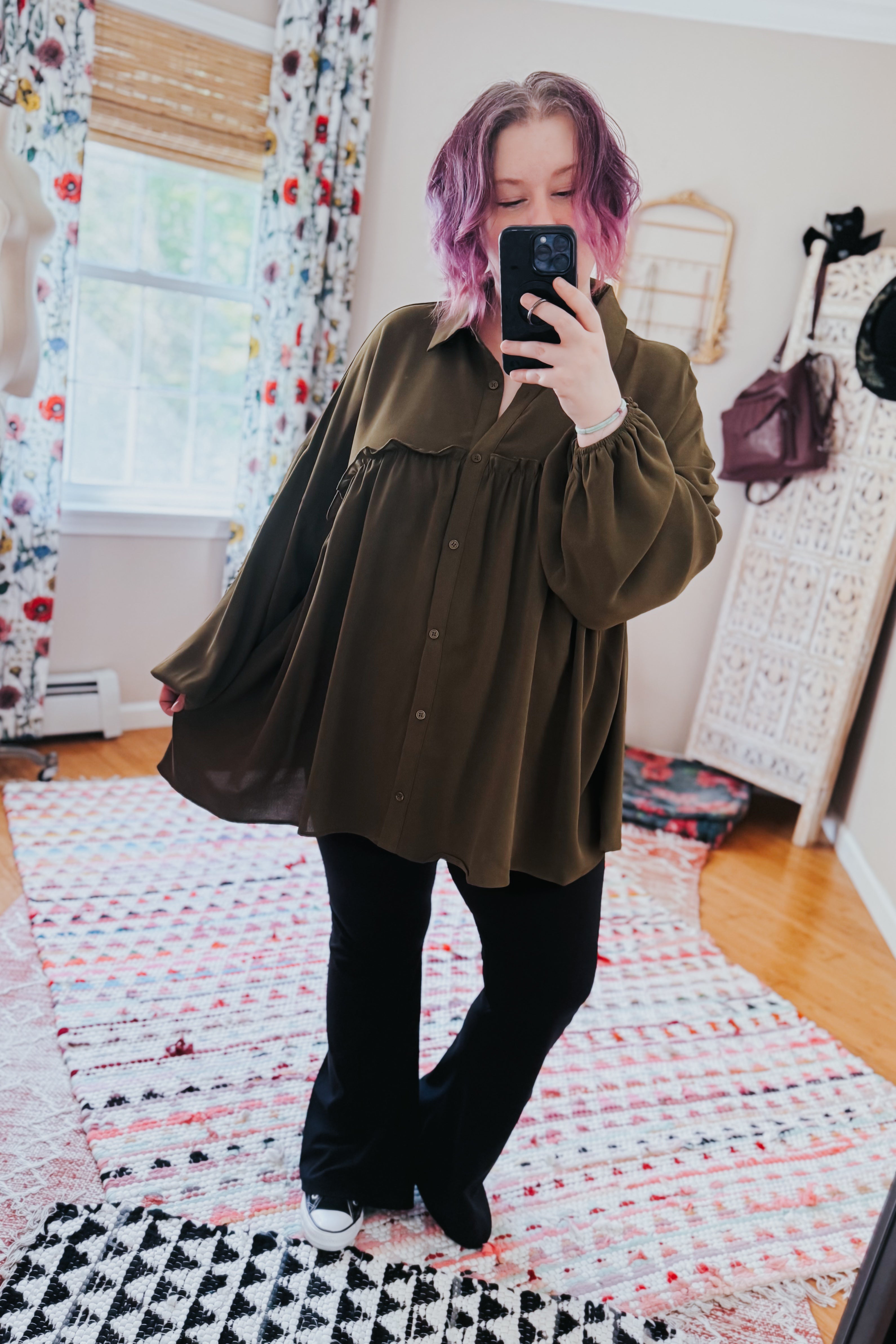 Woman with purple hair wearing loose olive button-down blouse and black pants taking mirror selfie indoors