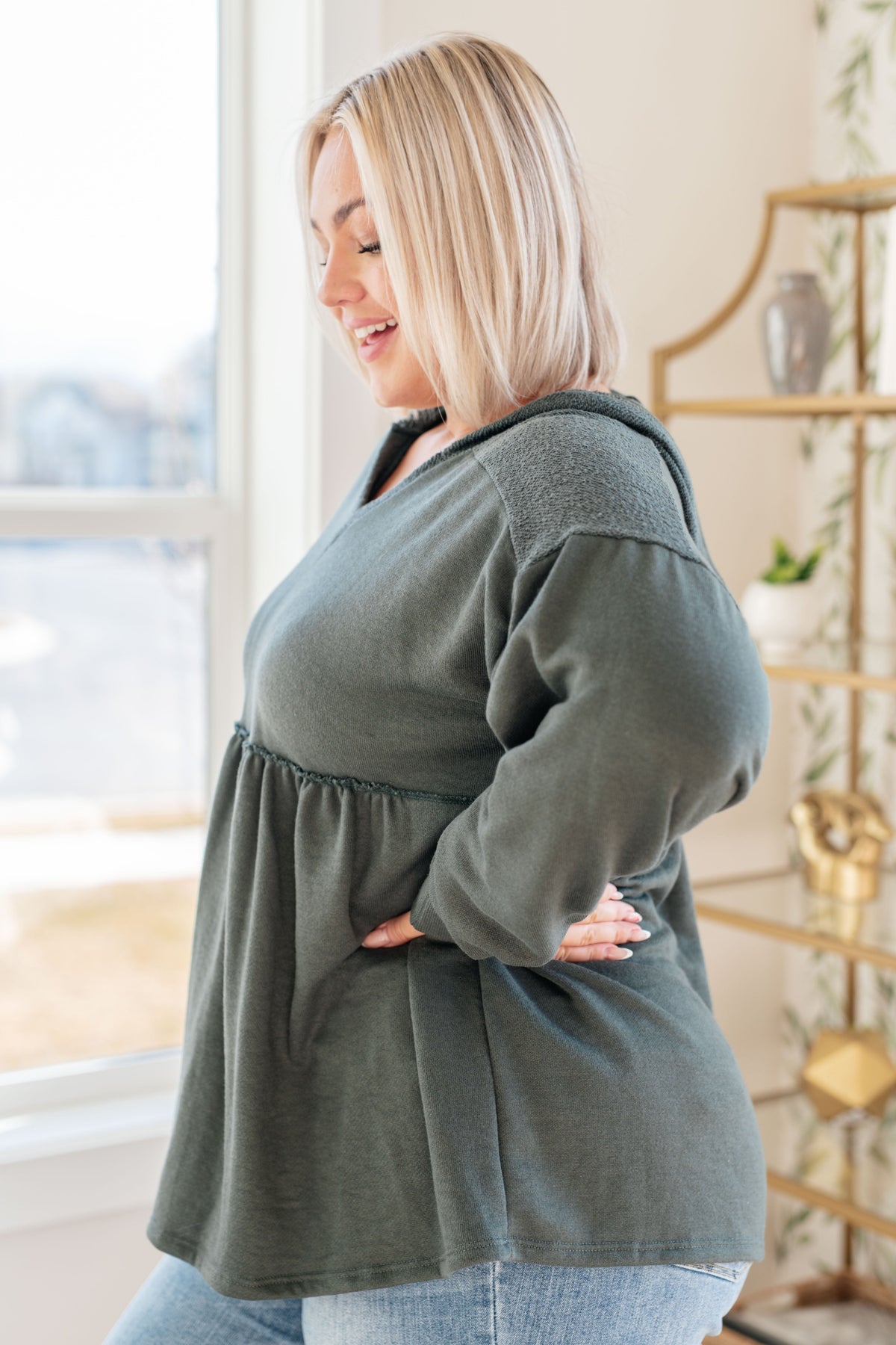 Woman modeling a teal hooded V neck pullover with gathered peplum and balloon sleeves over light wash jeans in a bright room