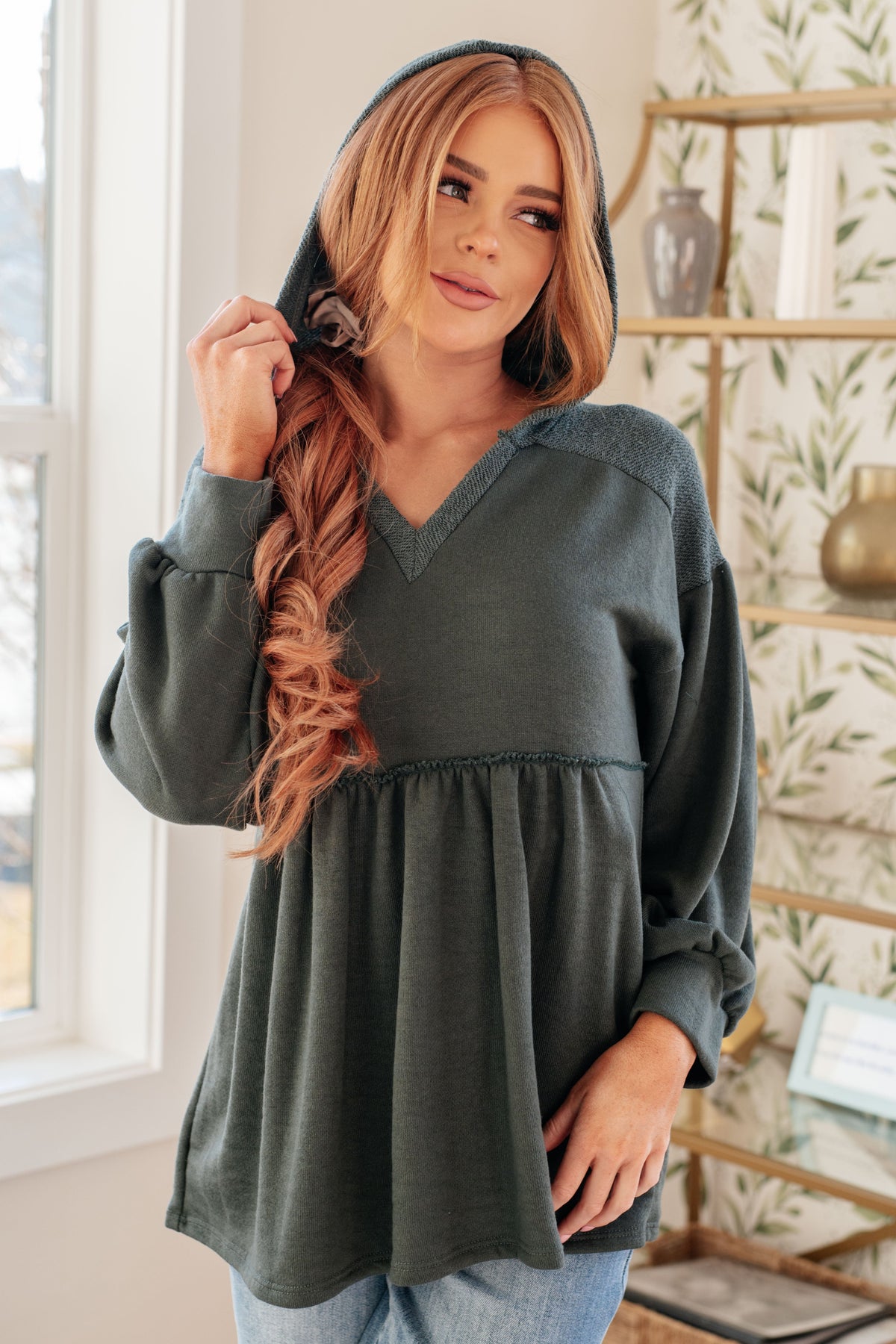 Woman wearing a teal hooded V neck pullover with gathered peplum and balloon sleeves styled with jeans indoors