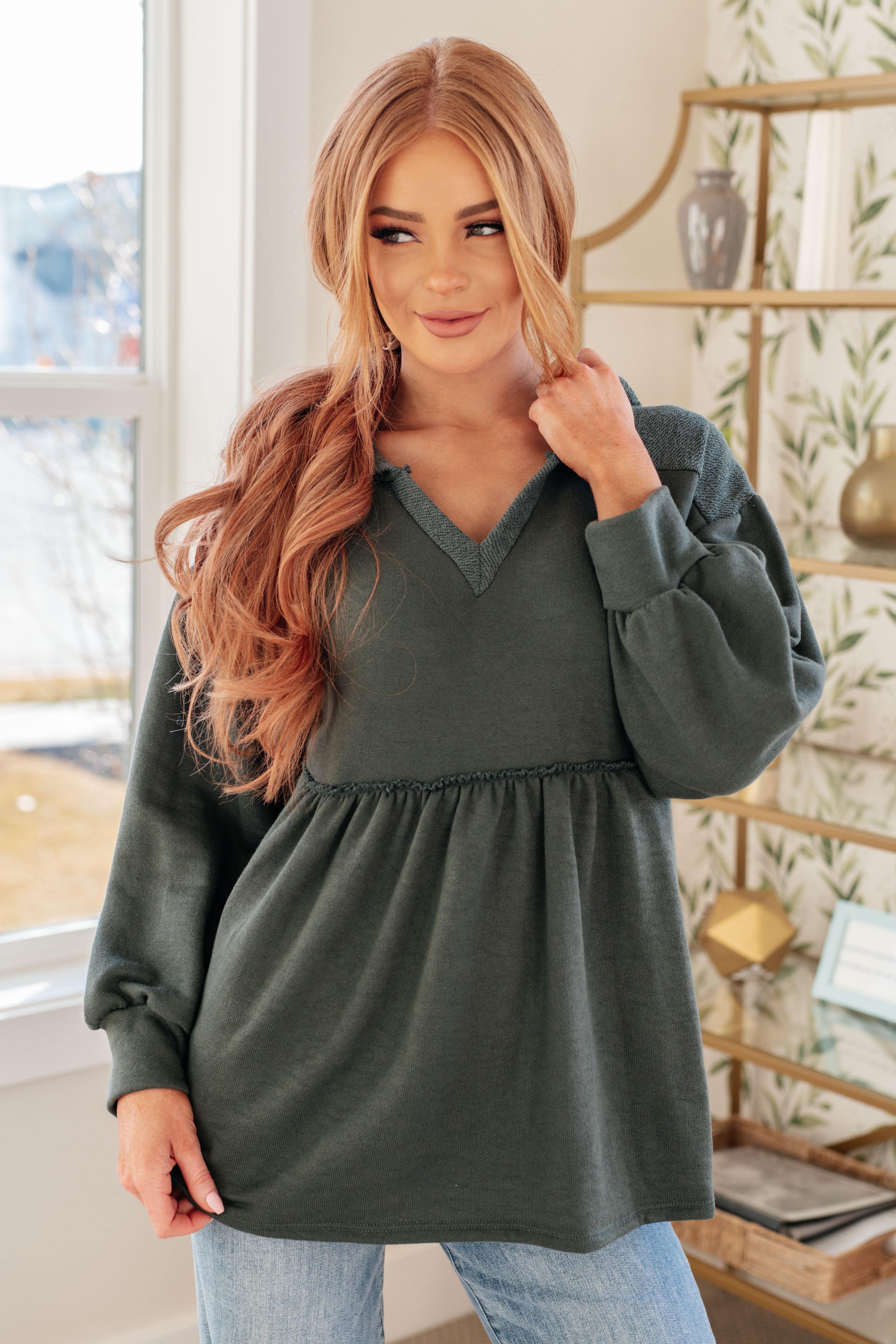 Woman wearing a dusty green v-neck hooded pullover and blue jeans in an indoor setting.