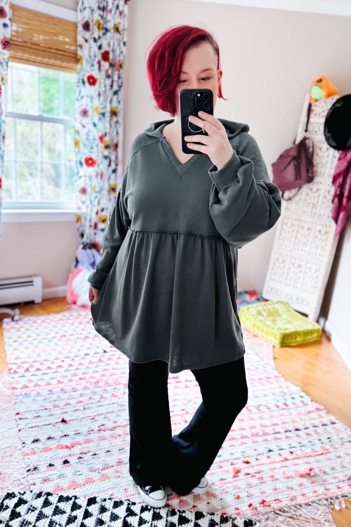 Woman taking a bedroom mirror selfie in a teal hooded V neck pullover with a gathered peplum and balloon sleeves black pants and white shoes