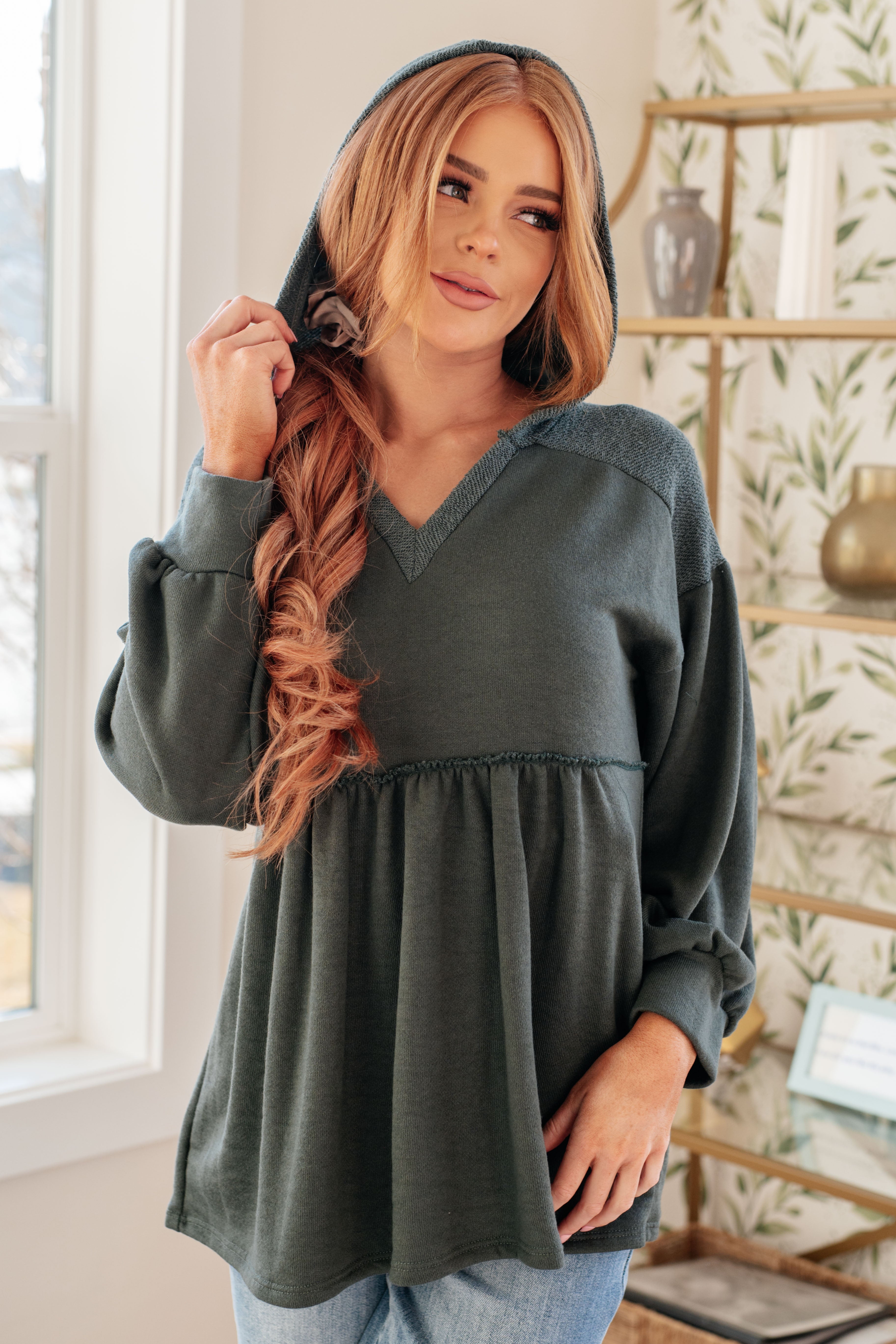 Woman wearing a dusty green v-neck hooded pullover and blue jeans in an indoor setting.