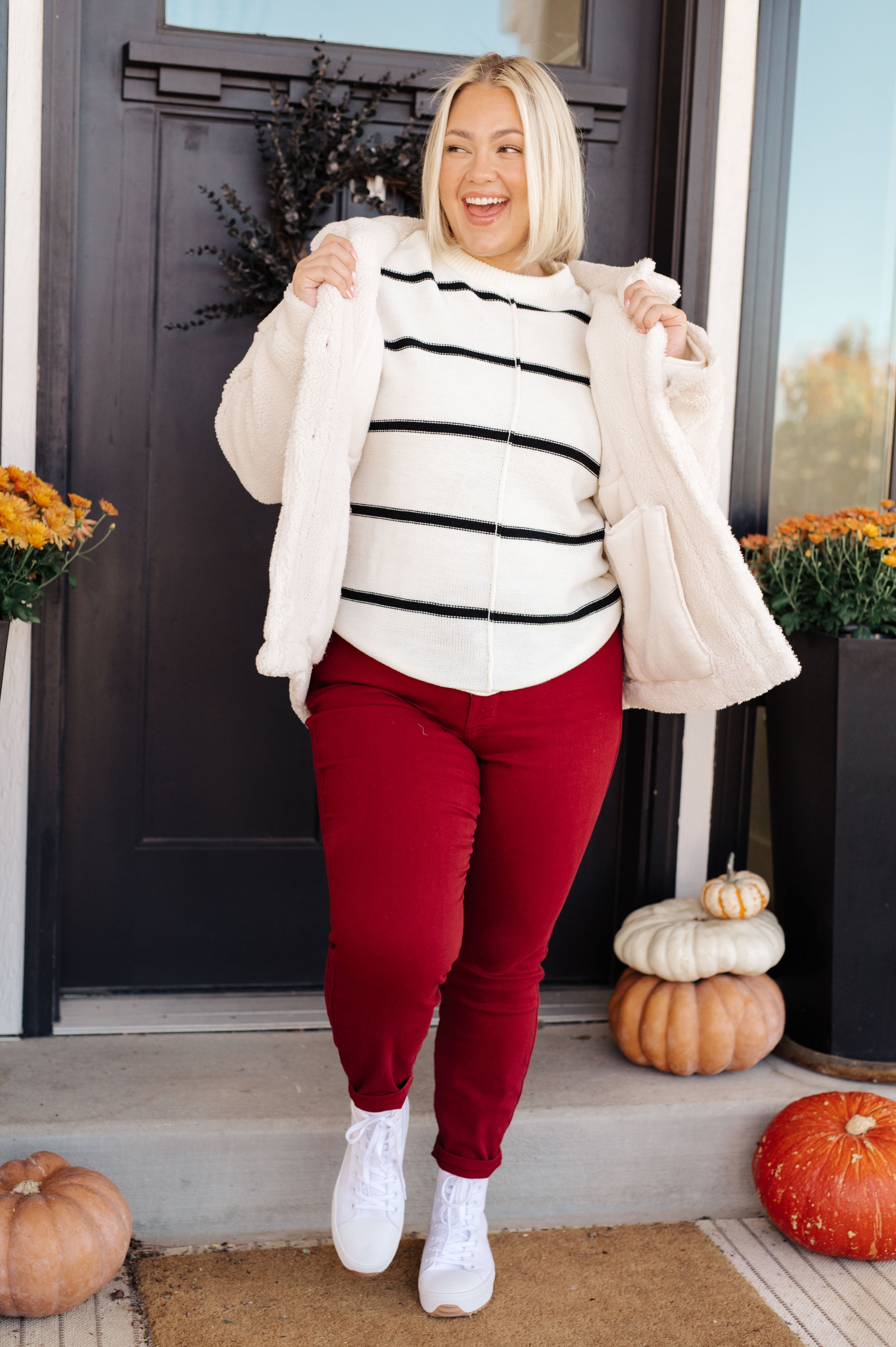 Woman smiling outside wearing white sherpa coat over striped sweater with red pants and white sneakers by pumpkins