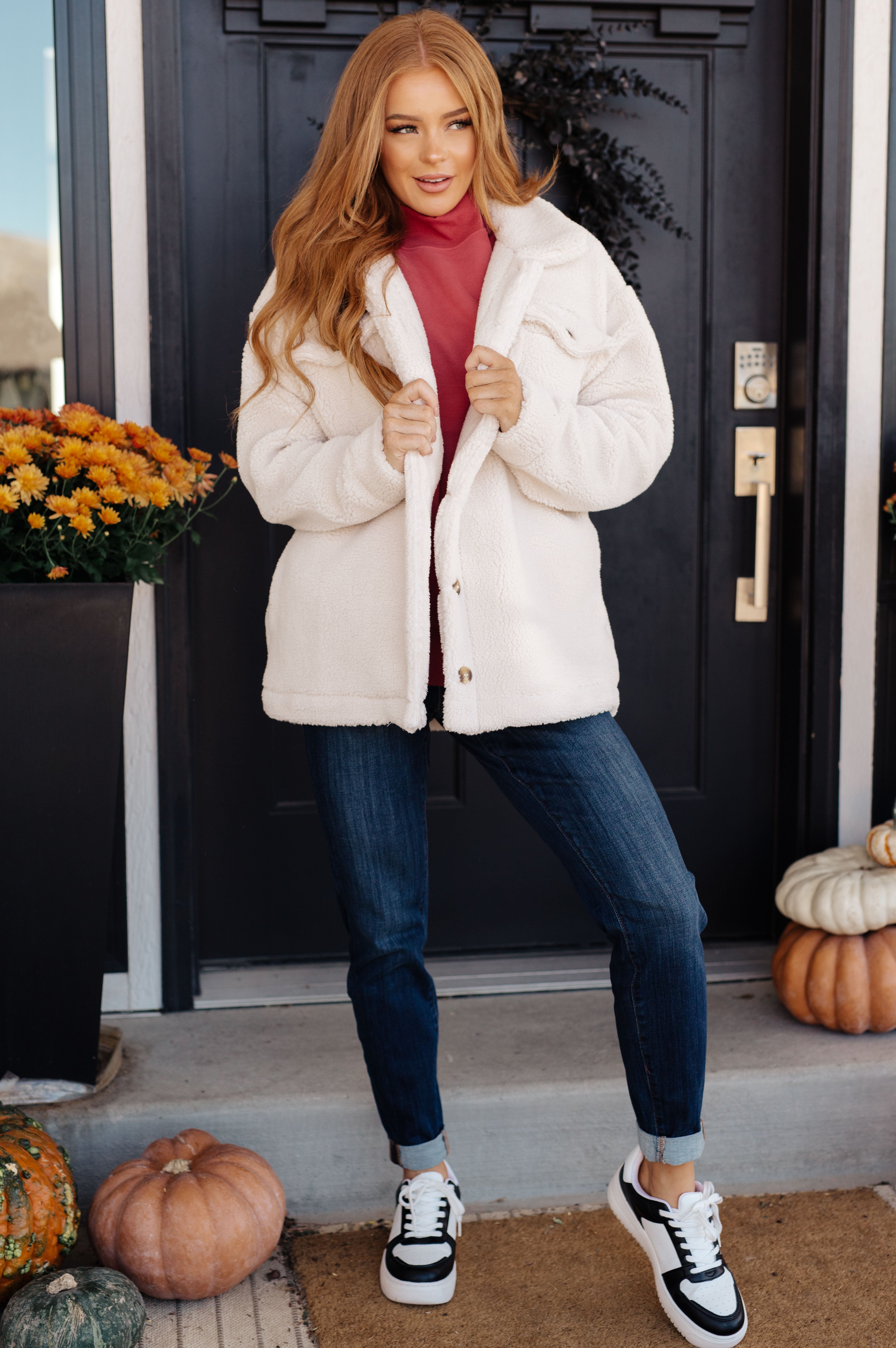 Woman wearing white plush sherpa coat over red top with dark denim jeans and black-and-white sneakers by front door with pumpkins