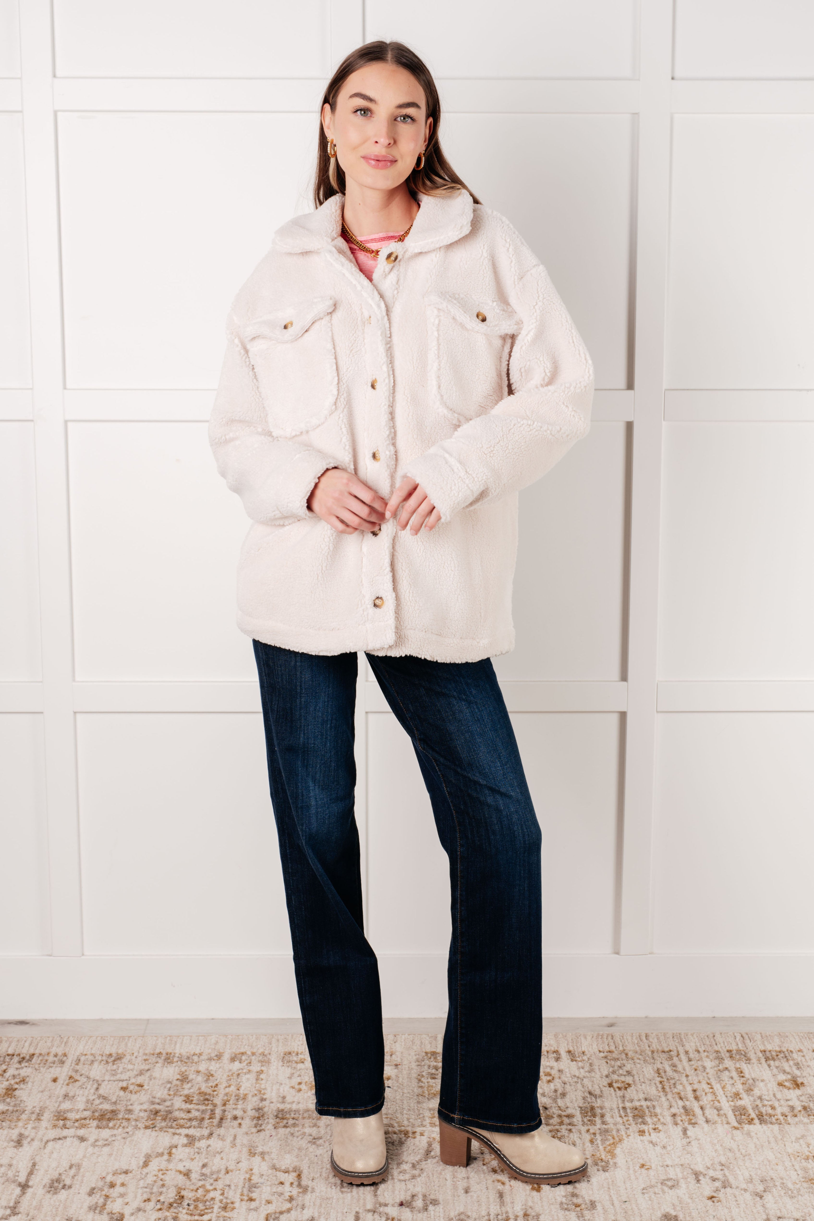 Woman wearing oversized white sherpa jacket with front pockets, dark jeans, and beige boots standing indoors