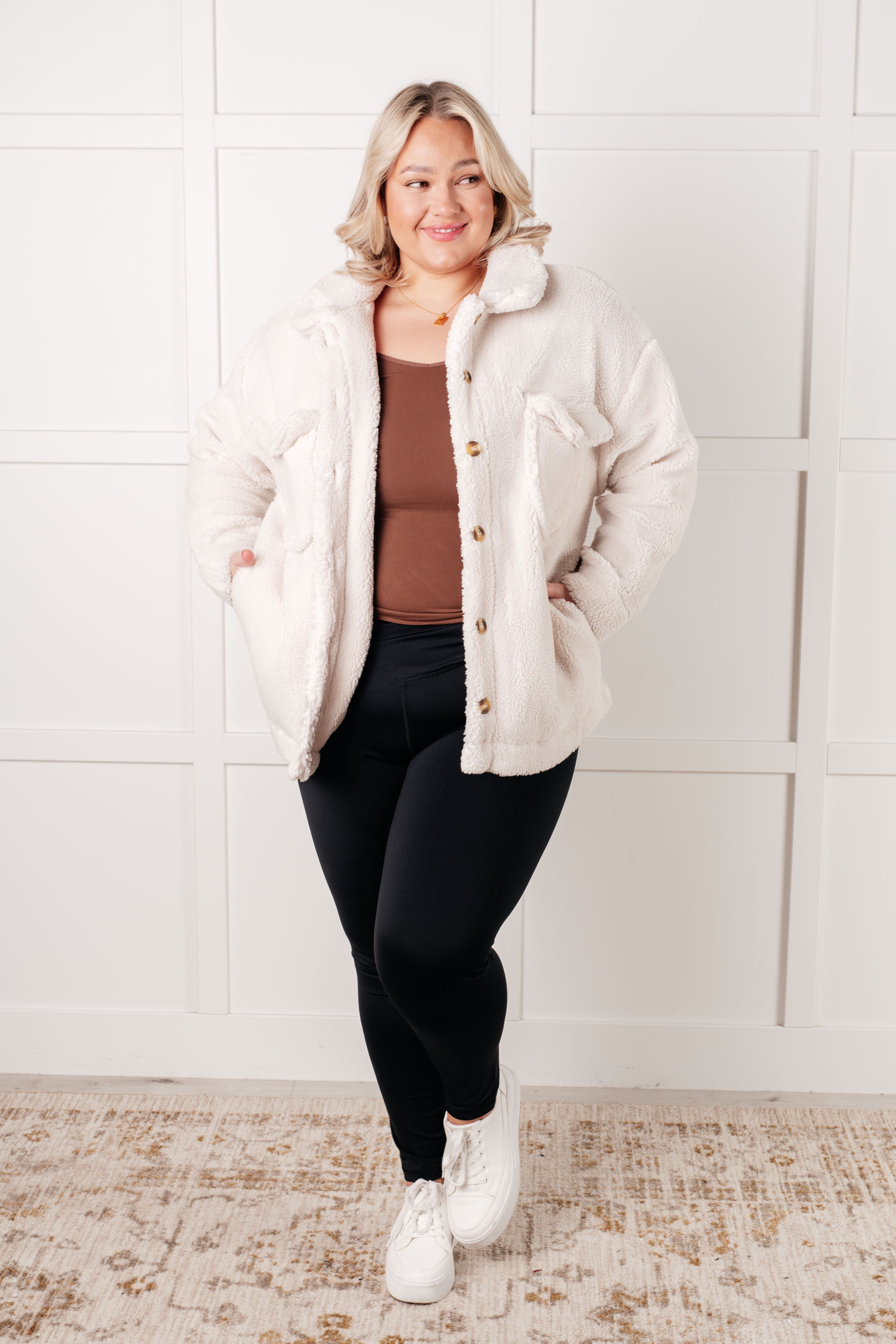 Smiling plus-size woman wearing white sherpa coat over brown top and black leggings standing in bright room