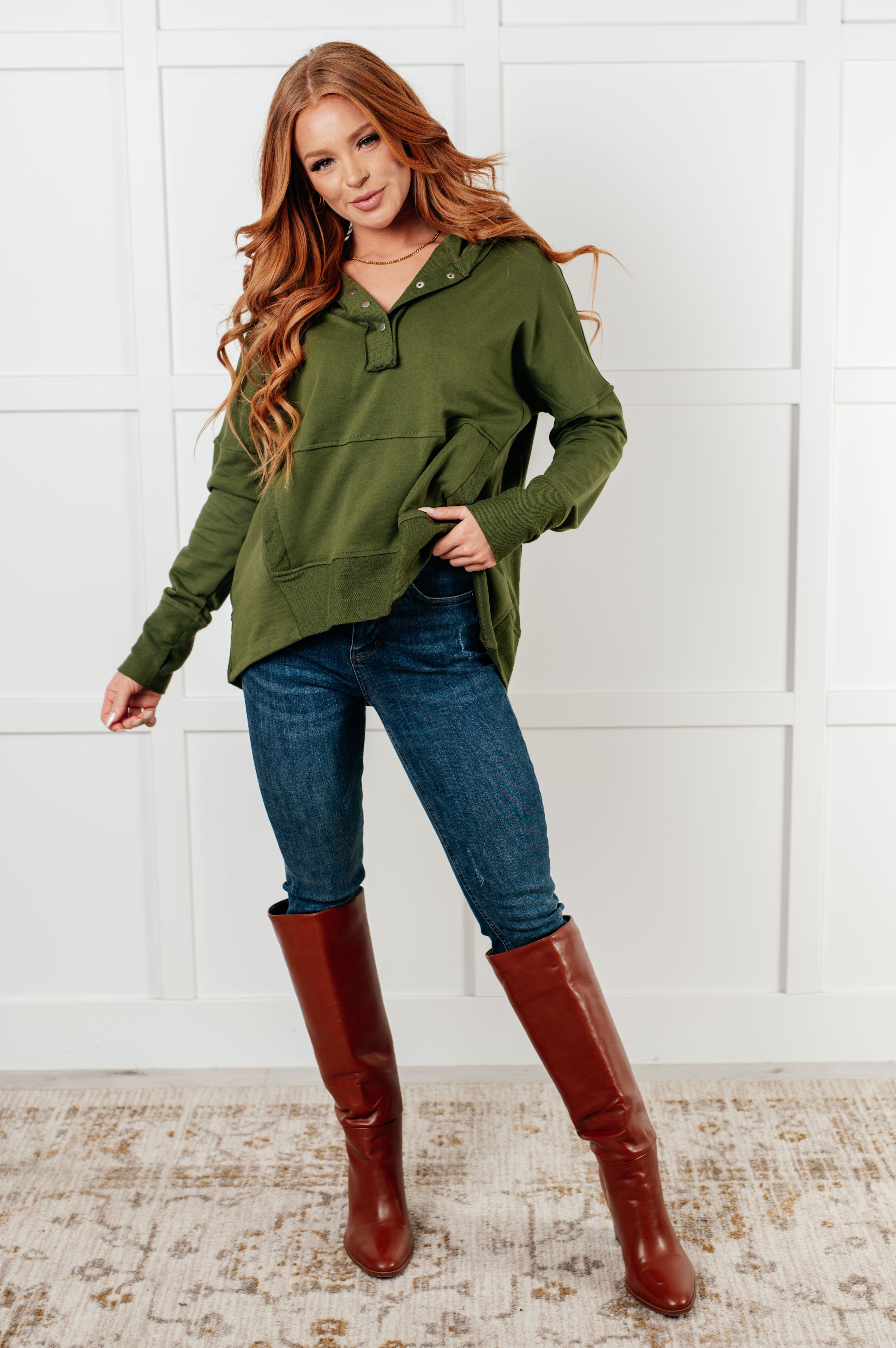 Woman wearing green snap hoodie with front pocket, dark jeans, and tall brown leather boots in bright studio setting