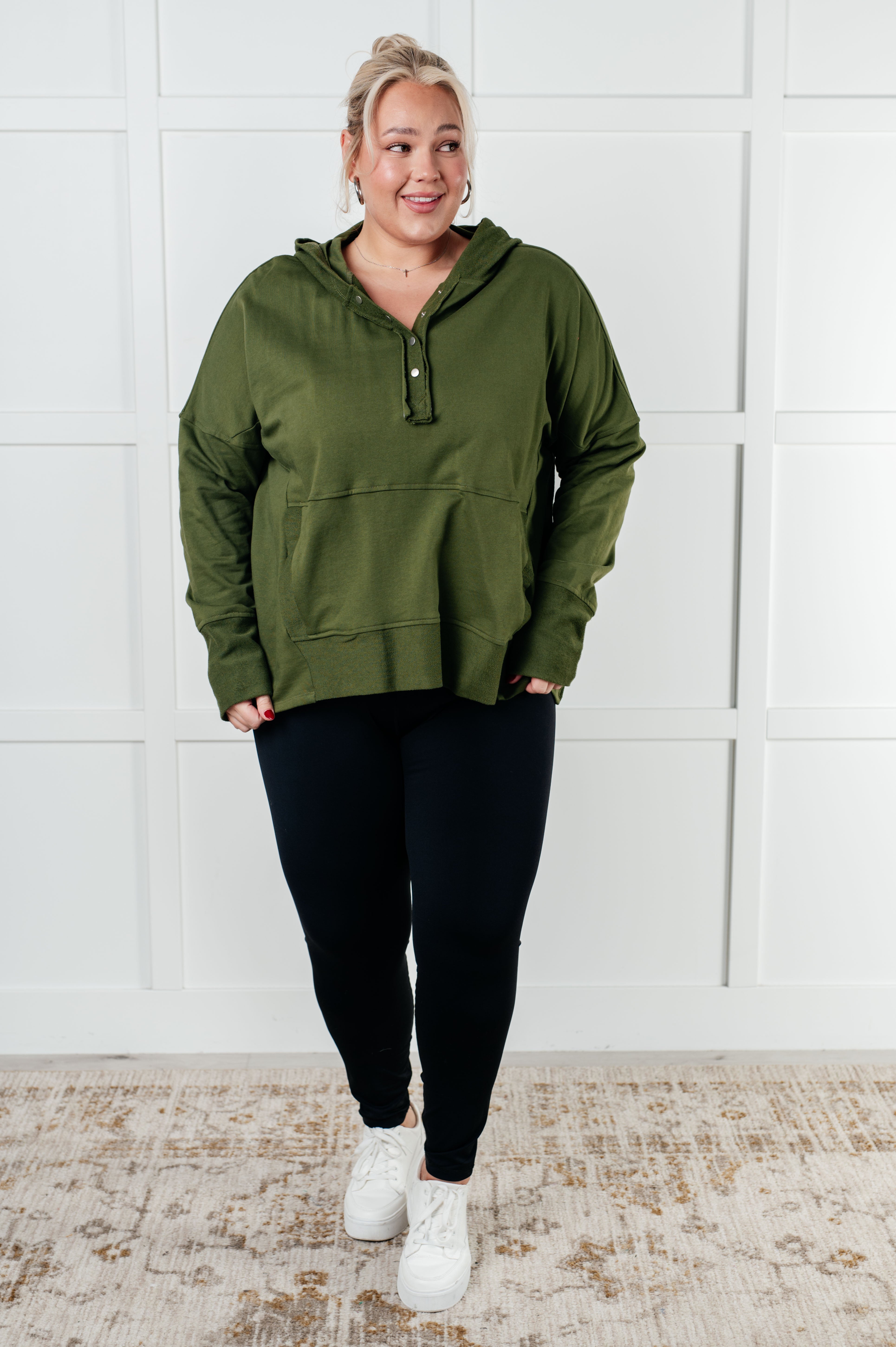 Smiling woman wearing olive green hooded pullover with snap buttons and black leggings standing on patterned rug