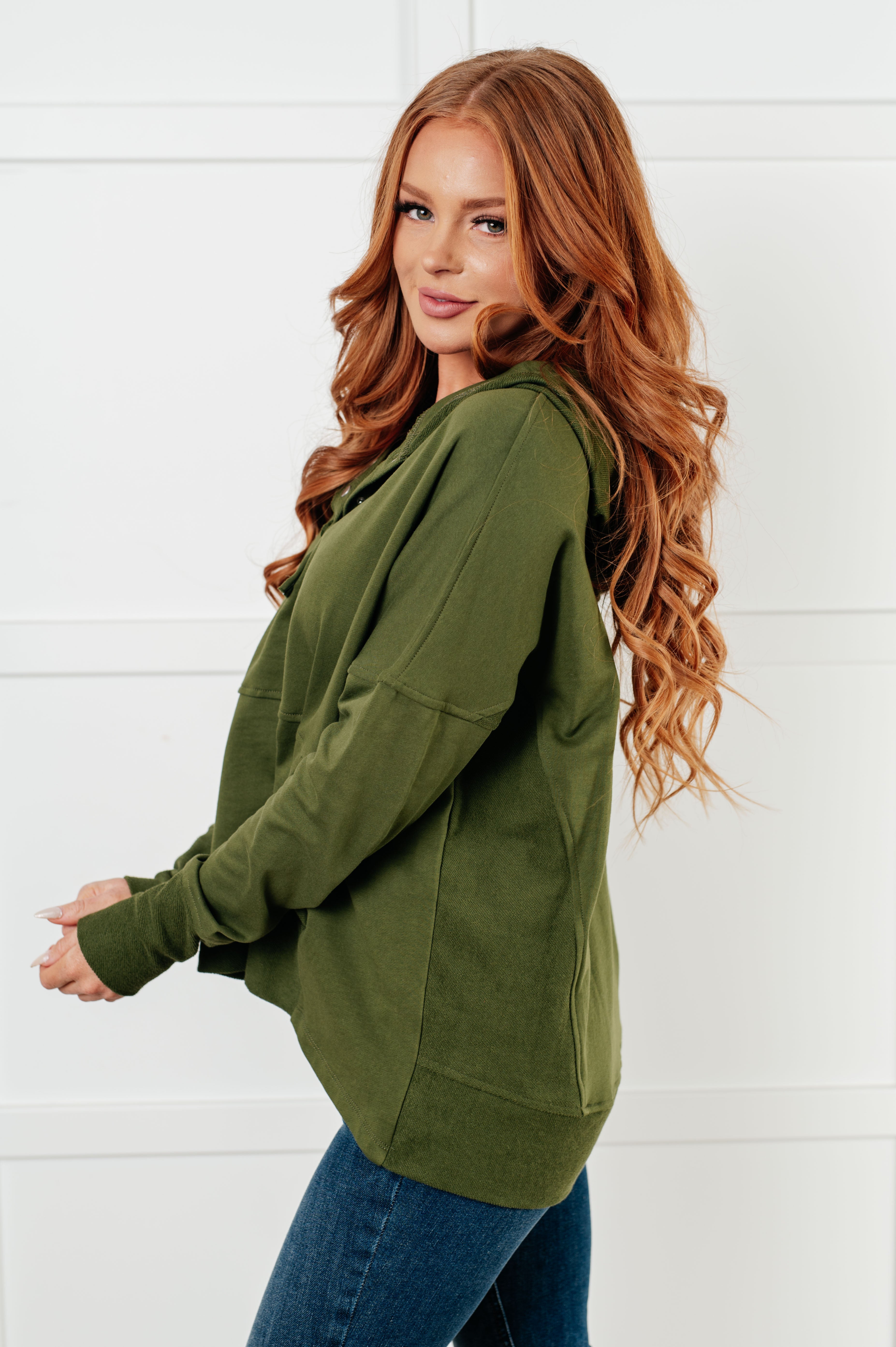 Woman with long red hair wearing relaxed green hooded pullover and blue jeans standing against white wall
