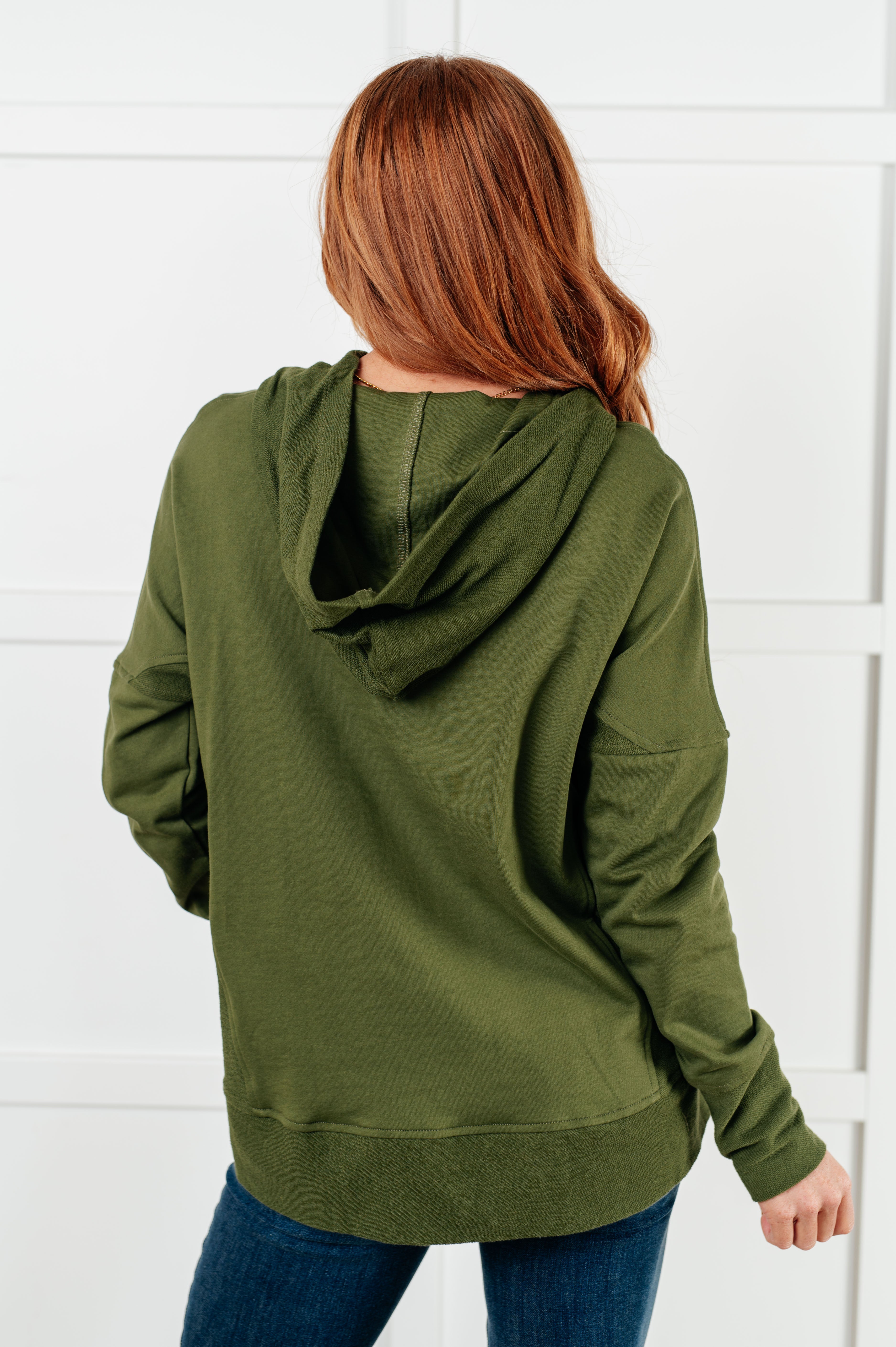 Woman shown from behind wearing relaxed-fit green hooded pullover with long sleeves and jeans in bright studio setting