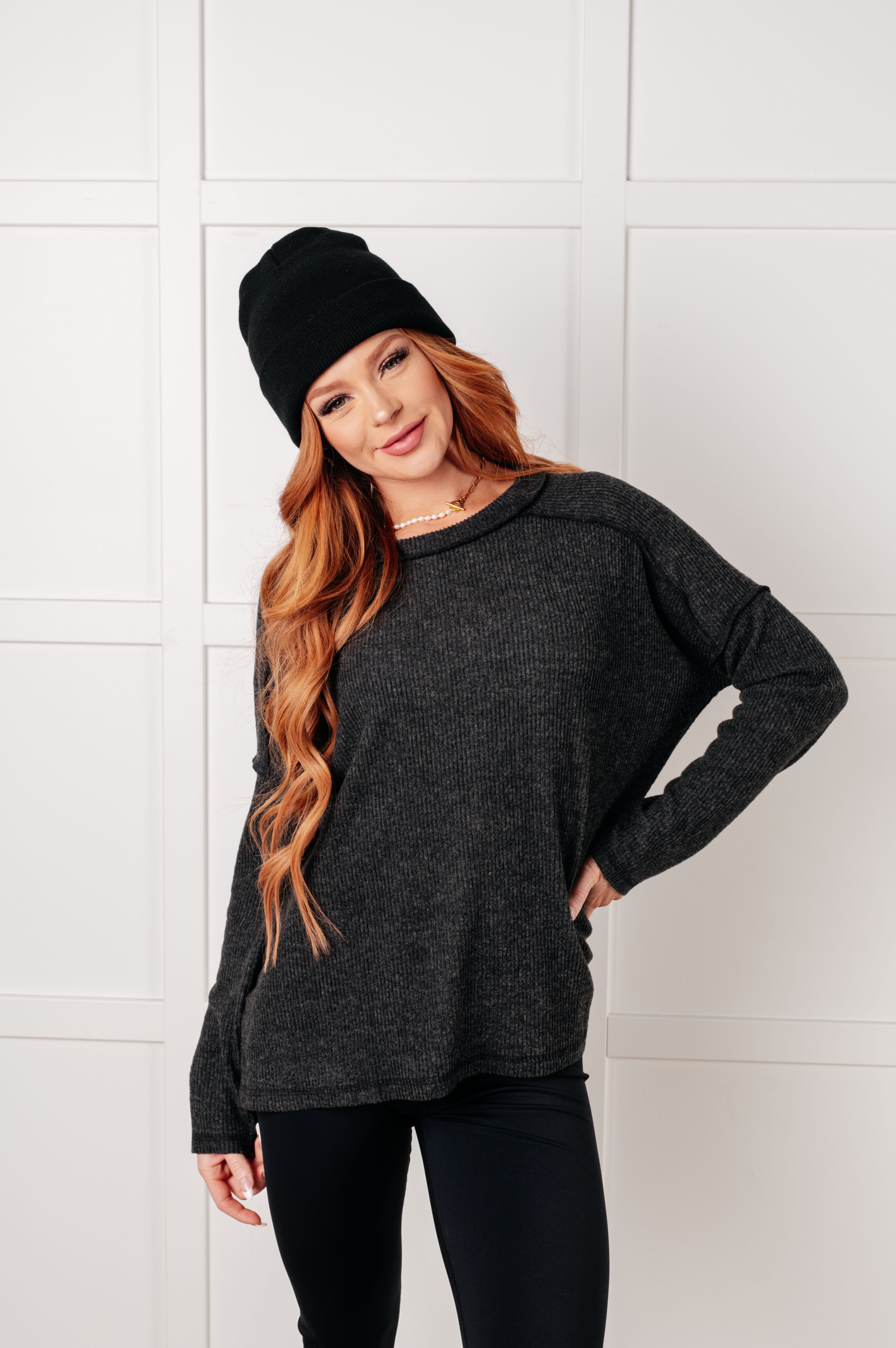 Woman with long red hair wearing relaxed black ribbed sweater and black beanie smiling in bright studio setting