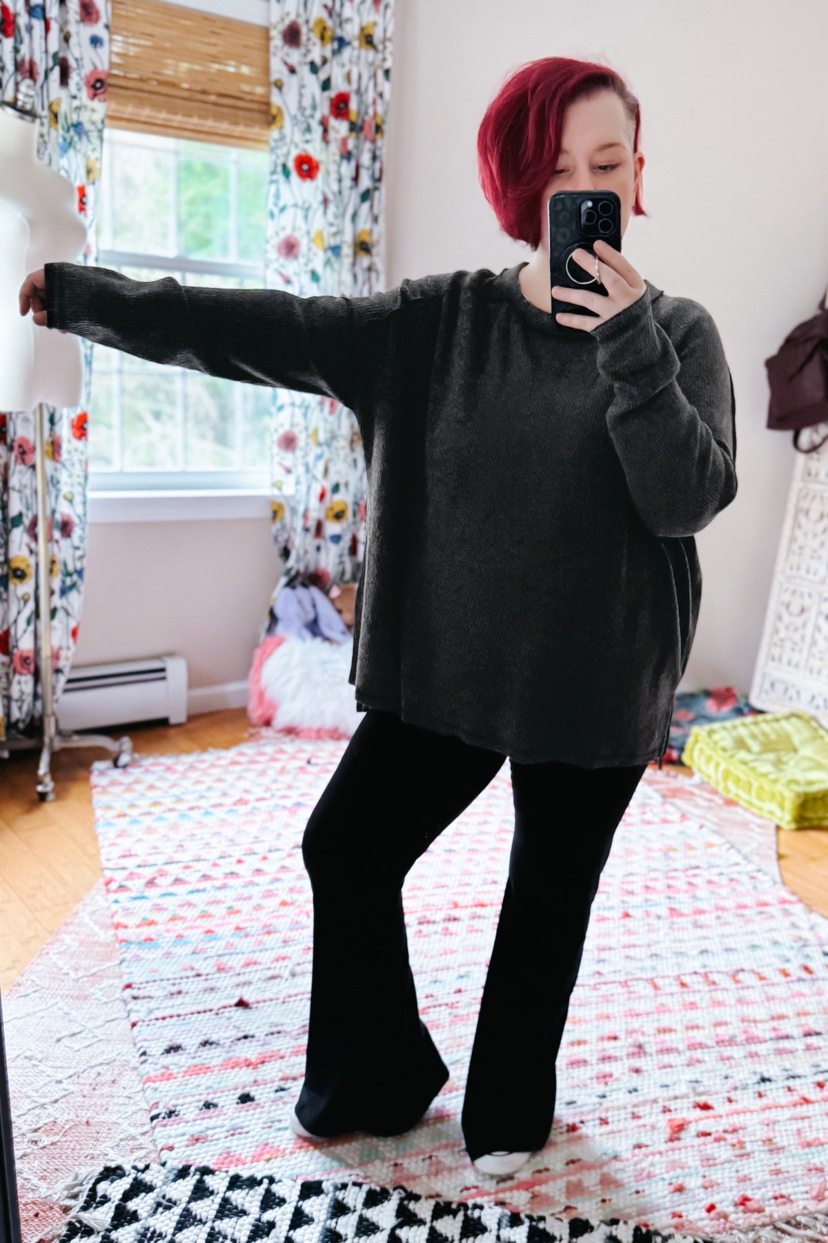 Red haired woman taking a bedroom mirror selfie in a black ribbed sweater and black flared pants on layered rugs