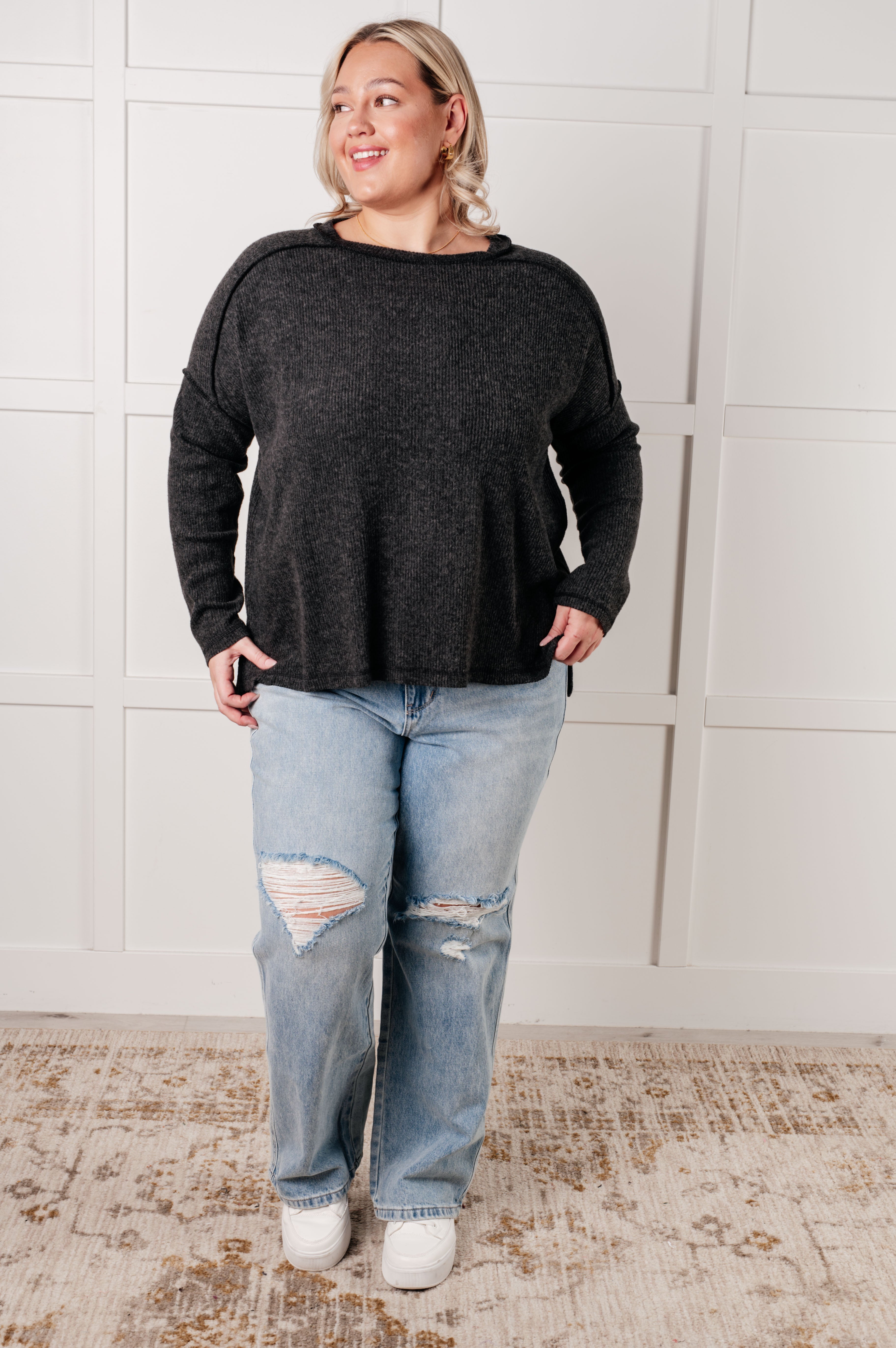 Plus-size woman wearing loose black ribbed sweater and distressed light blue jeans standing on patterned rug