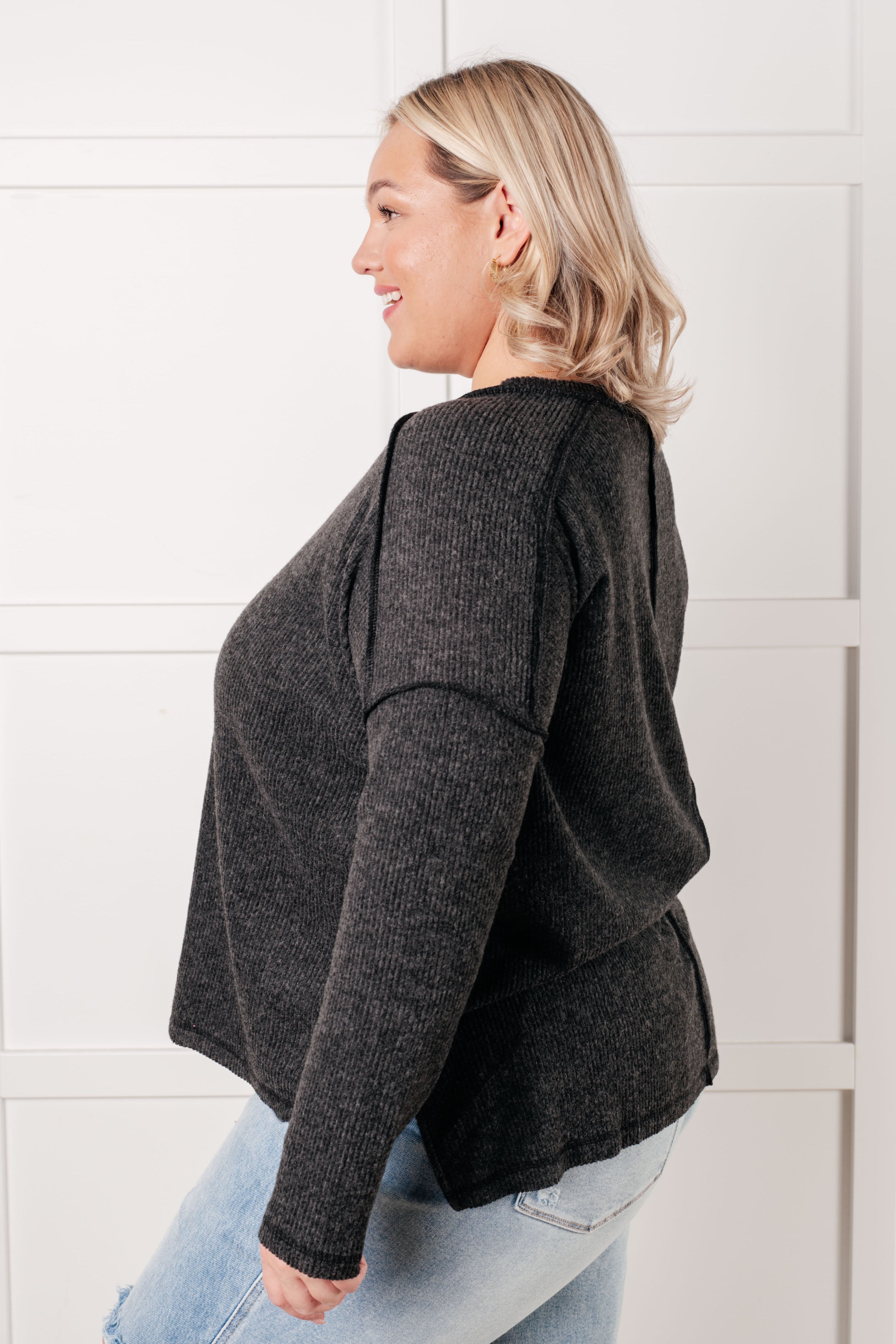 Side view of smiling woman wearing black ribbed sweater with long sleeves and relaxed fit paired with light-wash jeans