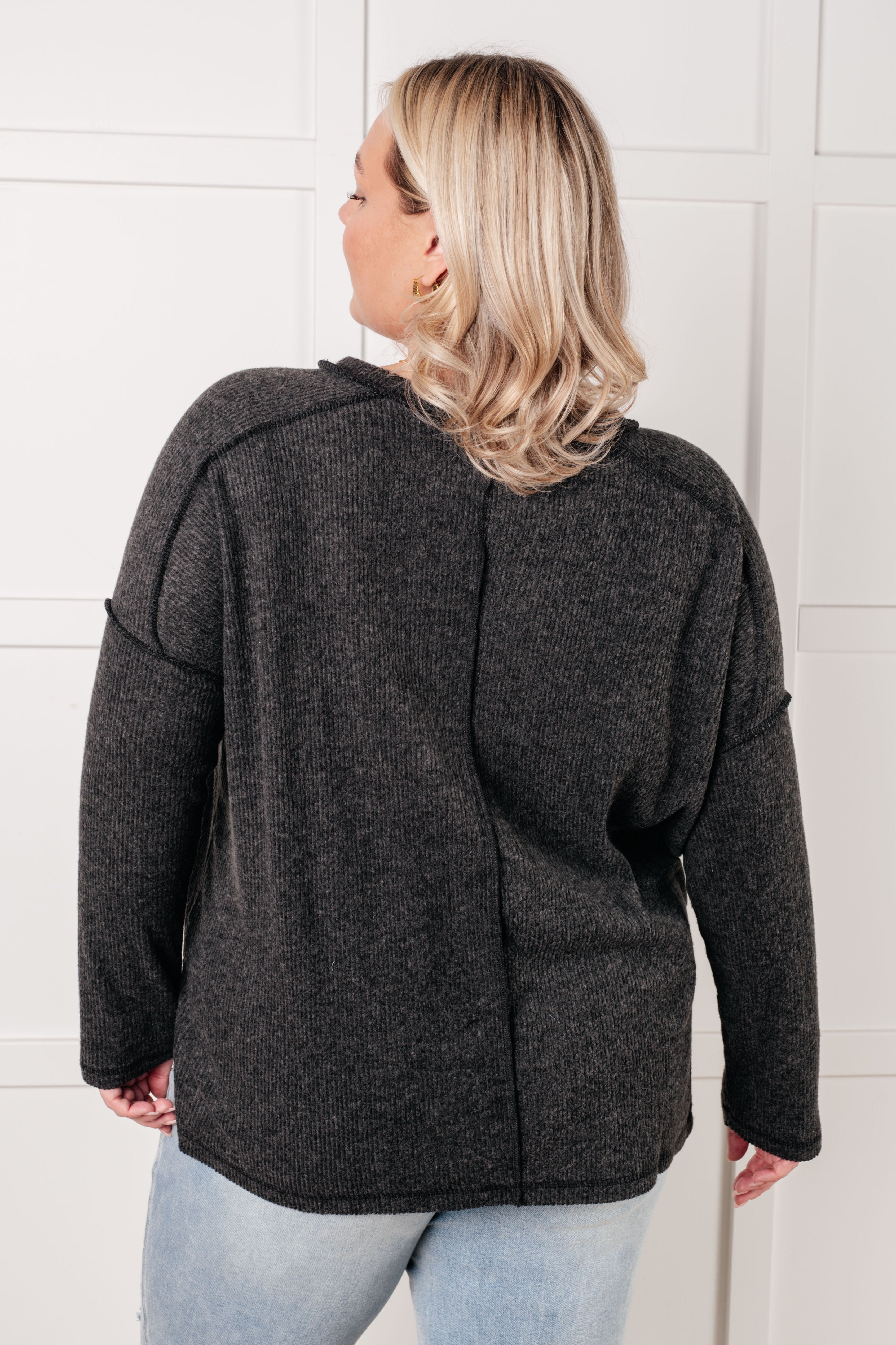 Woman wearing loose black ribbed sweater with long sleeves shown from behind against white wall