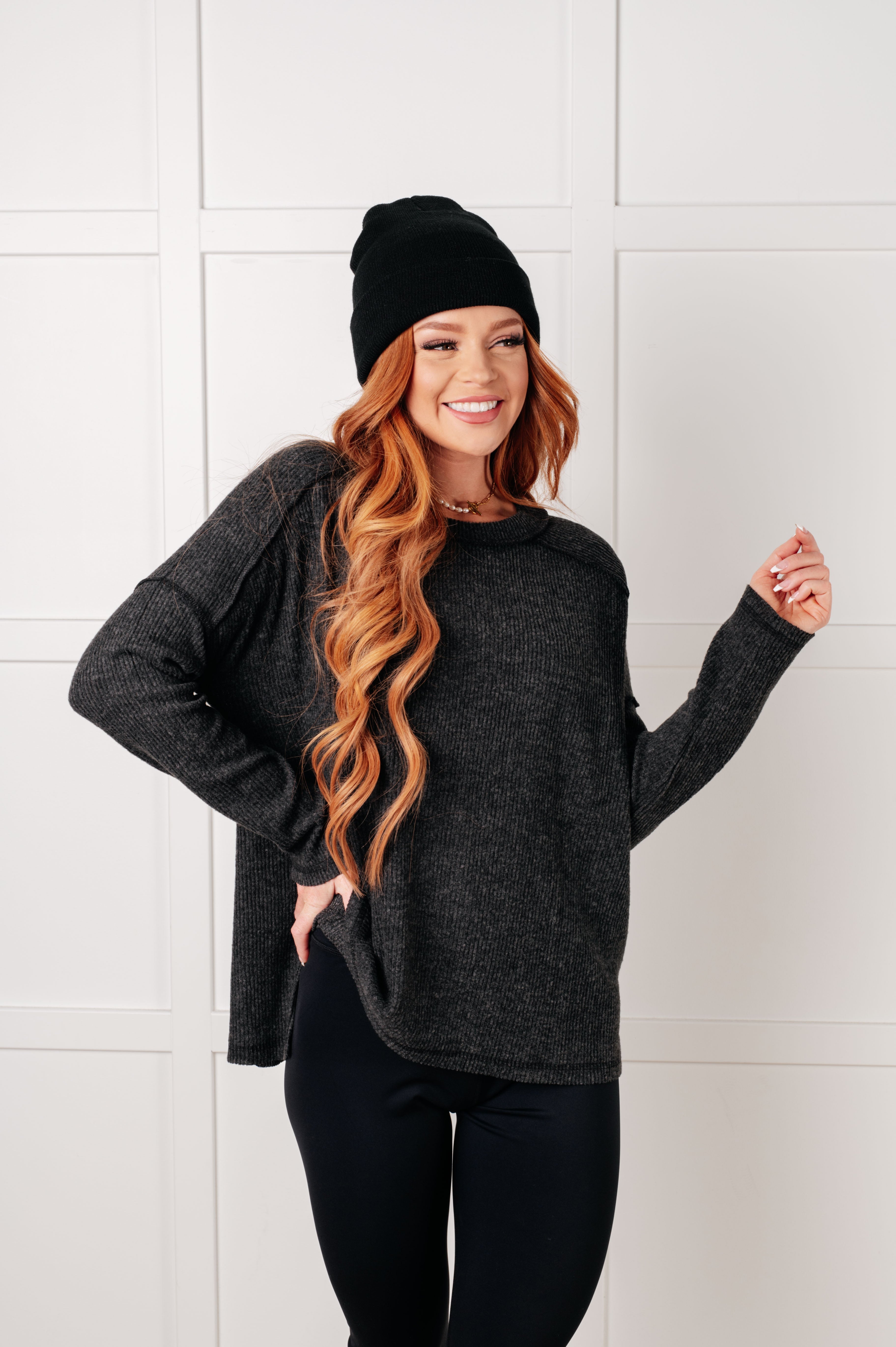 Smiling woman with long wavy hair wearing oversized black ribbed sweater and black beanie against light paneled wall