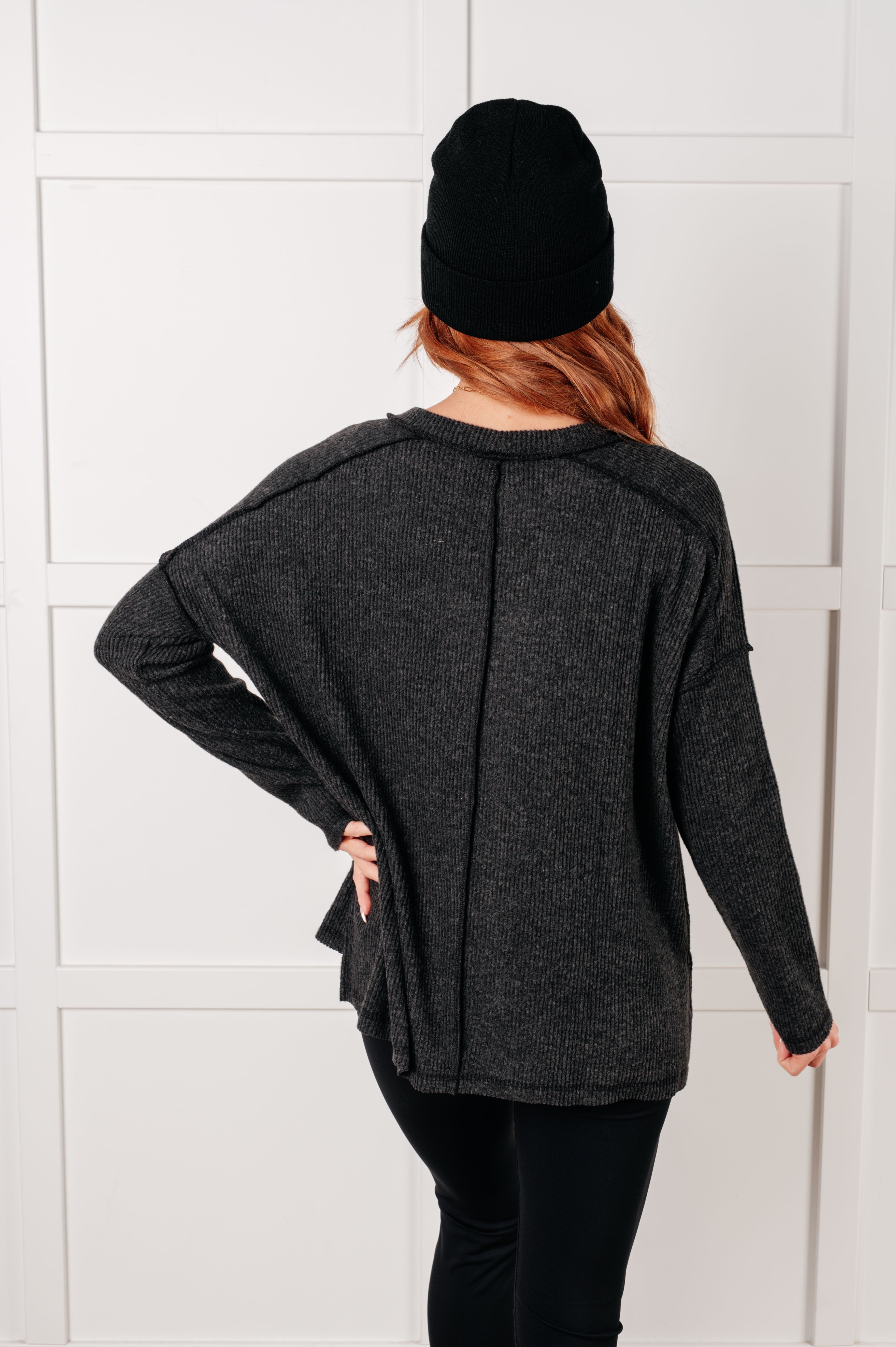 Woman seen from behind wearing loose black ribbed sweater and black beanie in simple white interior setting