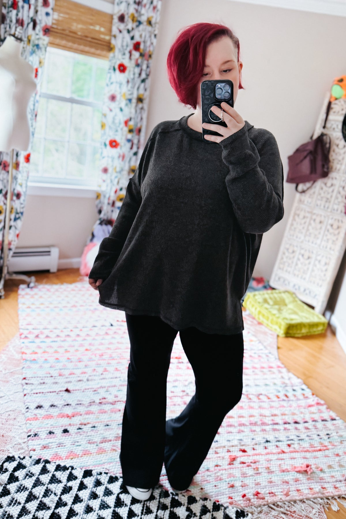 Red haired woman taking a mirror selfie in a black ribbed sweater and black pants over layered colorful rugs