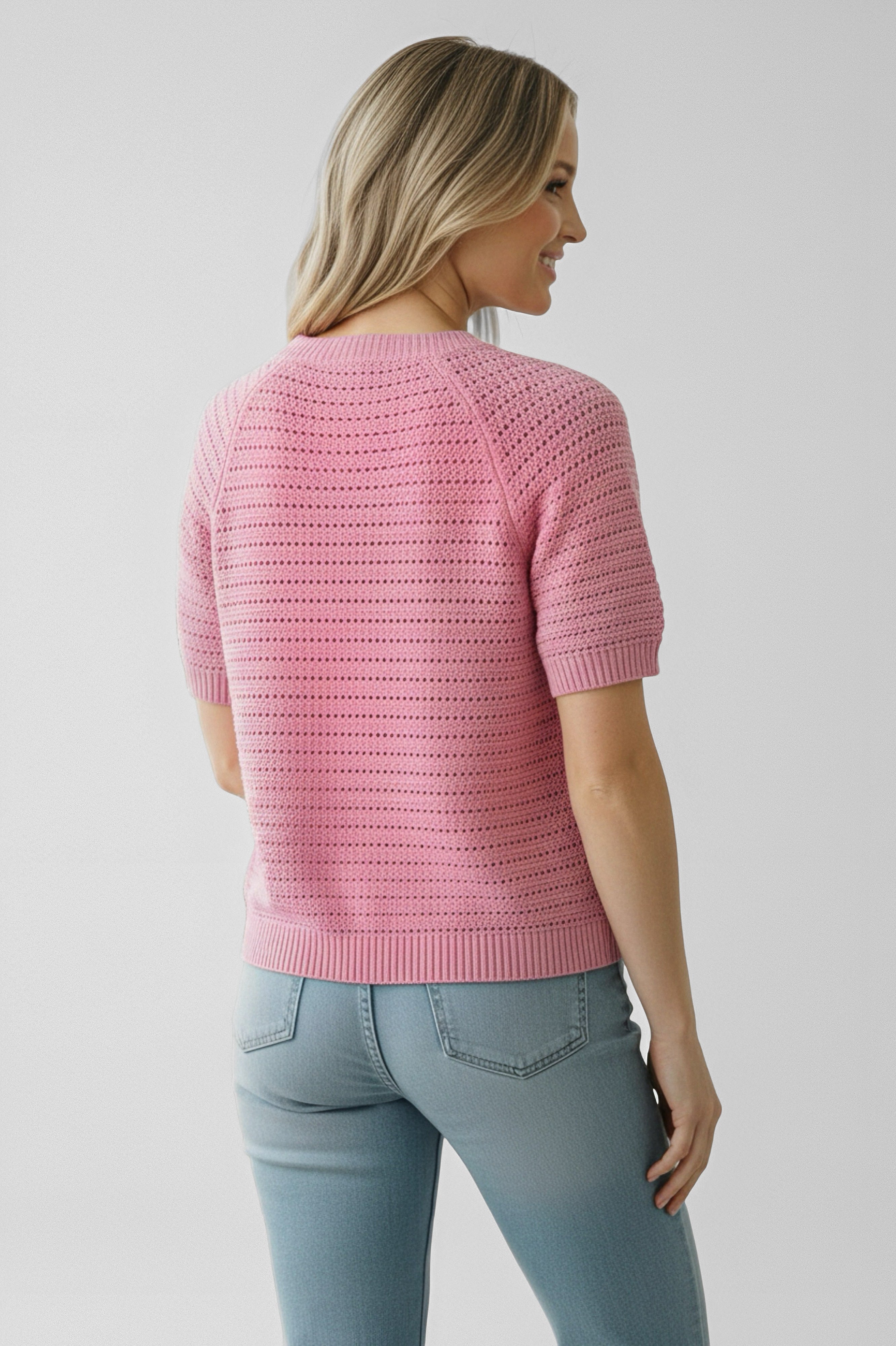 Woman with blonde hair wearing a pink short-sleeve knit top and light blue jeans standing with her back turned